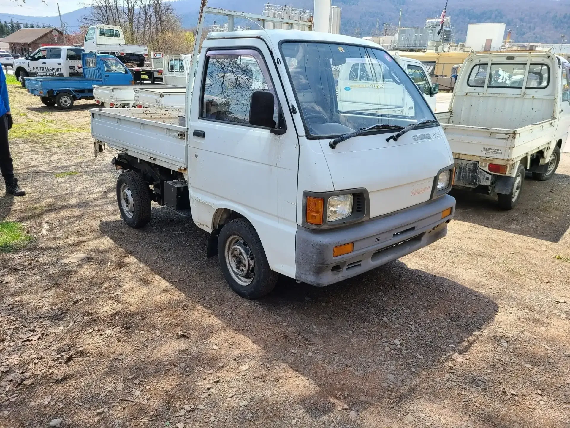 A white, used kei truck parked on a gravel lot, with another white truck partially visible in the background.
