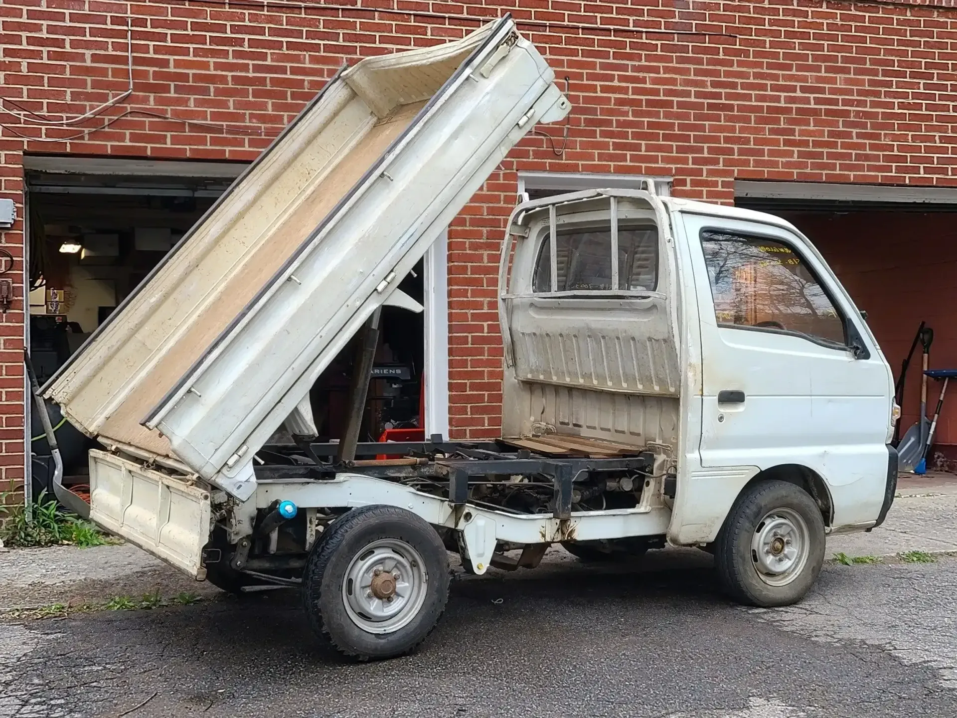 A small, white dump truck parked on gravel in front of a red brick building with its truck bed raised.