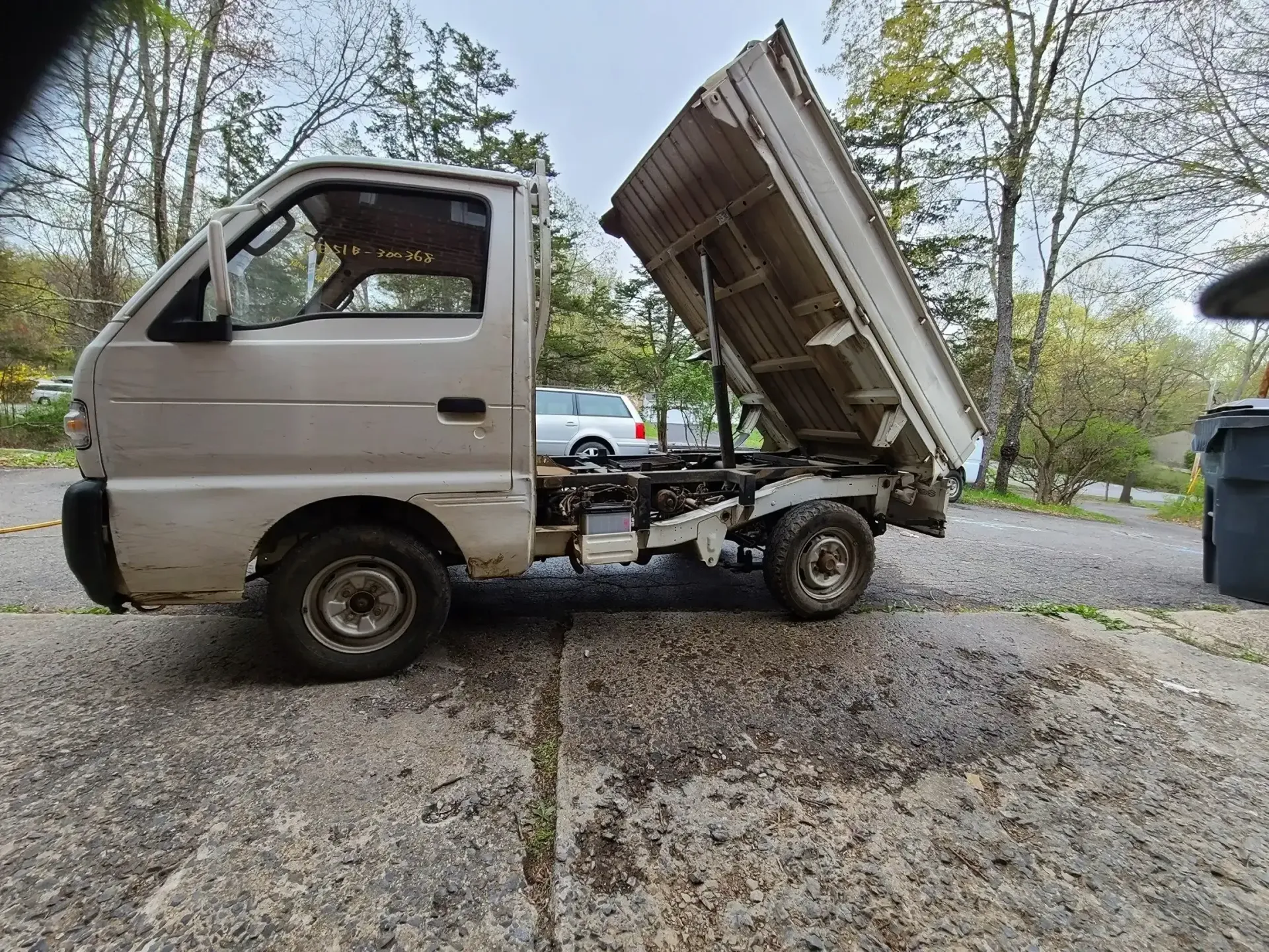 A light-colored, compact dump truck parked on a gravel driveway with its bed raised in the air.