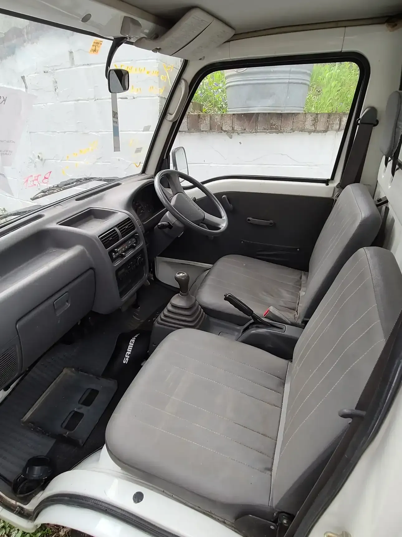 Interior view of a right-hand drive small truck with grey fabric seats, a manual gear stick, and a simple dashboard.