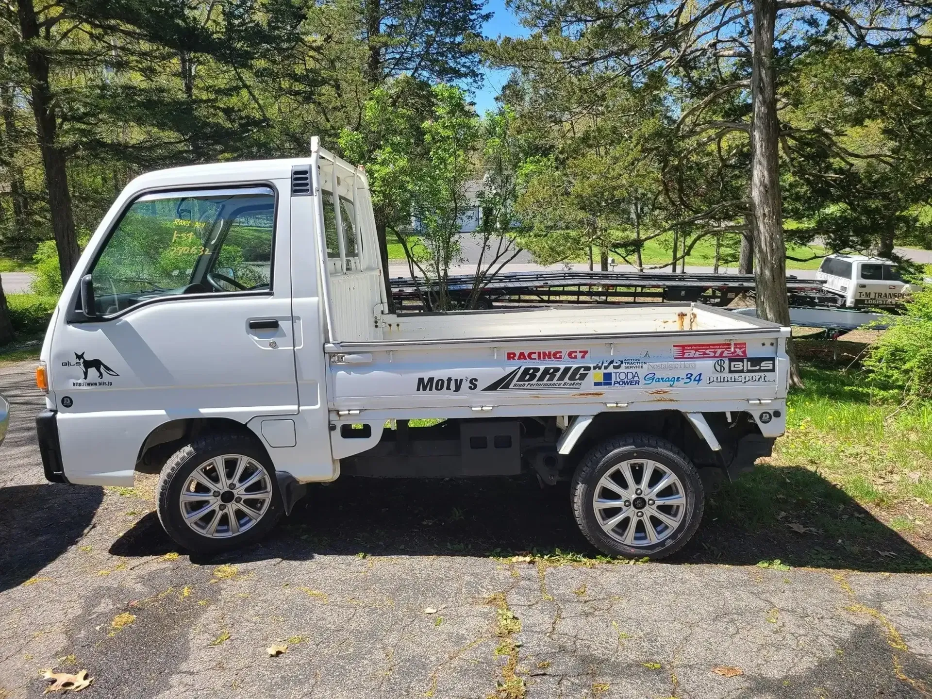 A white kei truck with custom aftermarket wheels parked on a gravel surface outdoors with trees in the background.