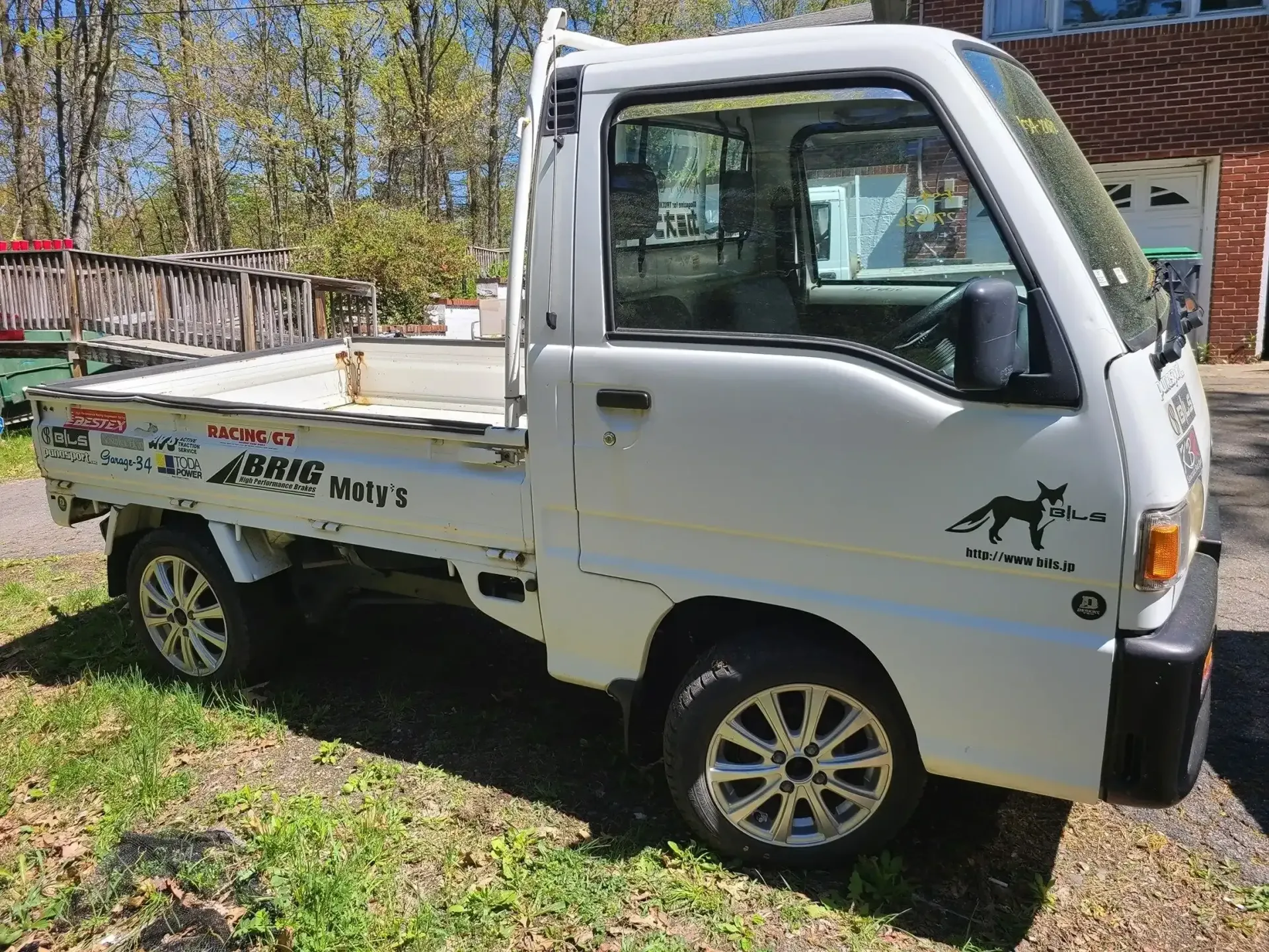 A white mini-truck parked on grass next to a house, featuring custom alloy wheels and decals on the side panels.
