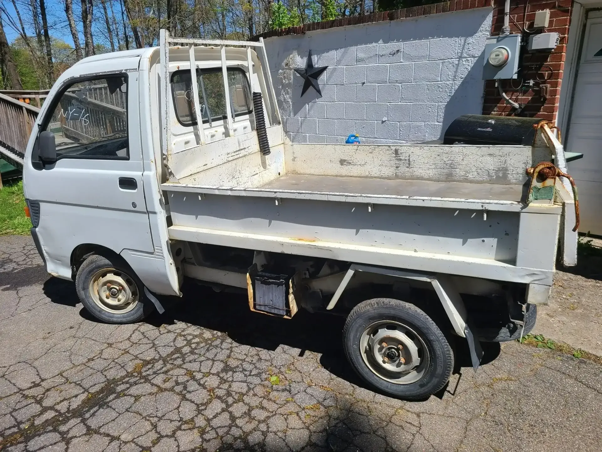 A white, older-model Japanese Kei pickup truck parked on a paved driveway in front of a white wall with a star.