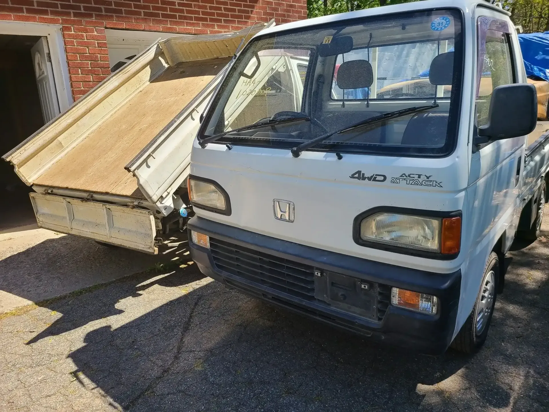 A white Honda Acty mini dump truck parked on a gravel surface next to a brick building.