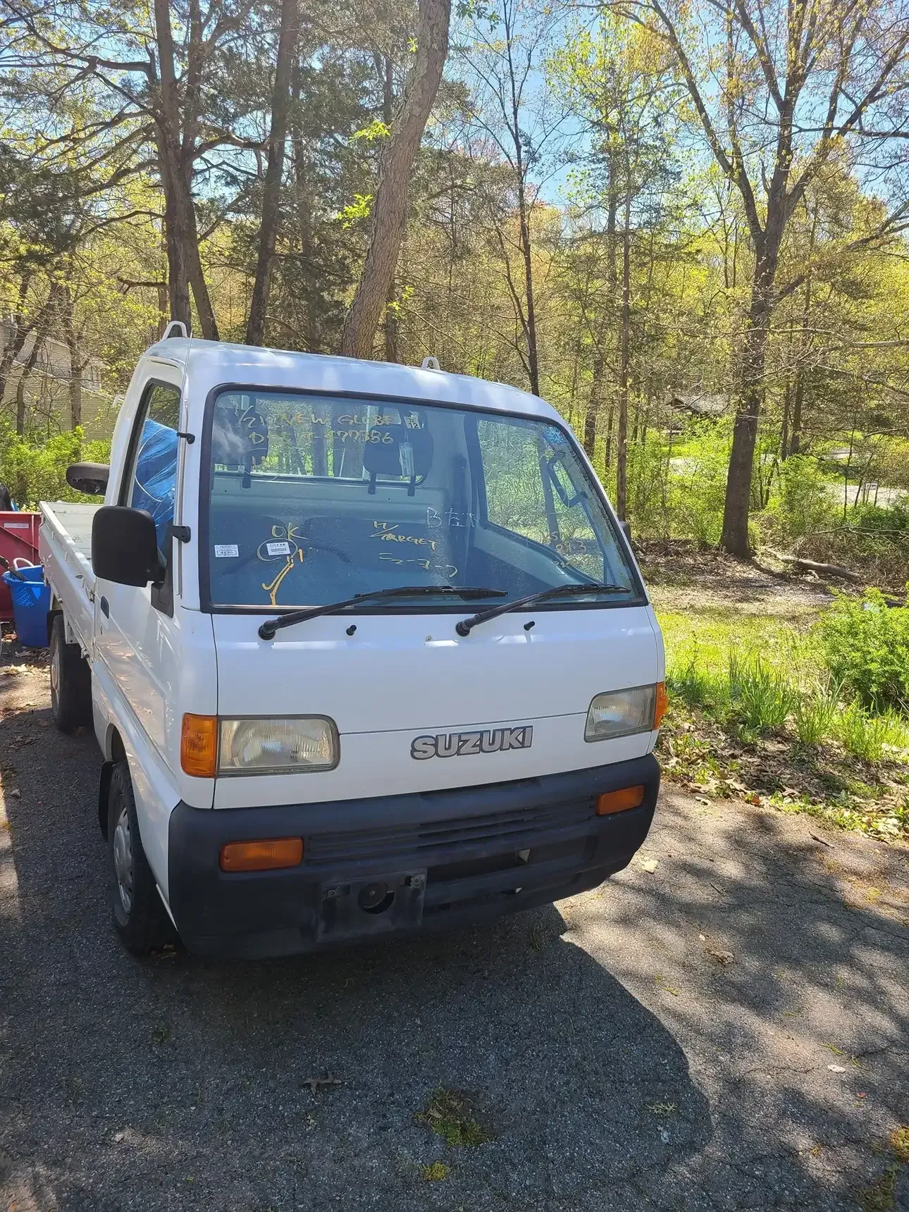A white Suzuki kei truck parked on a gravel surface outdoors with trees in the background.