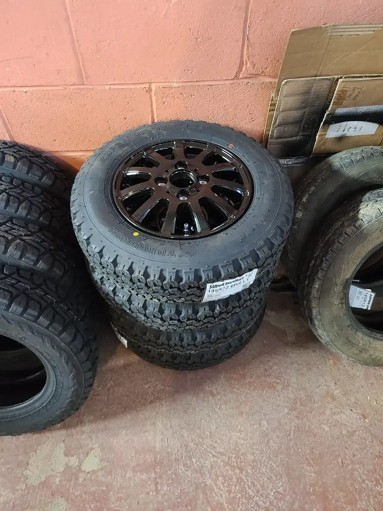 A stack of black off-road tires with black rims sitting on a concrete floor next to a cardboard box.