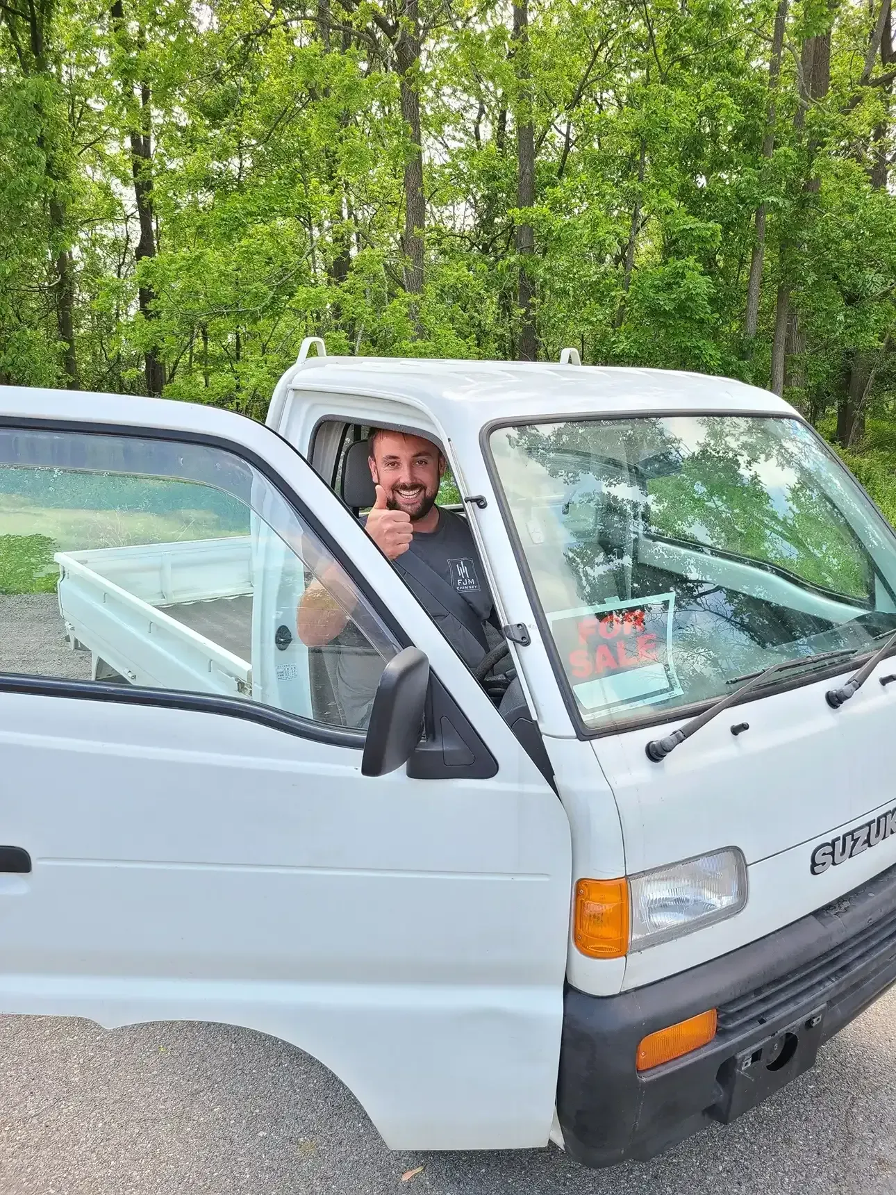 A smiling person in a white micro-truck gives a thumbs up from the driver's seat with the door open against a forest.