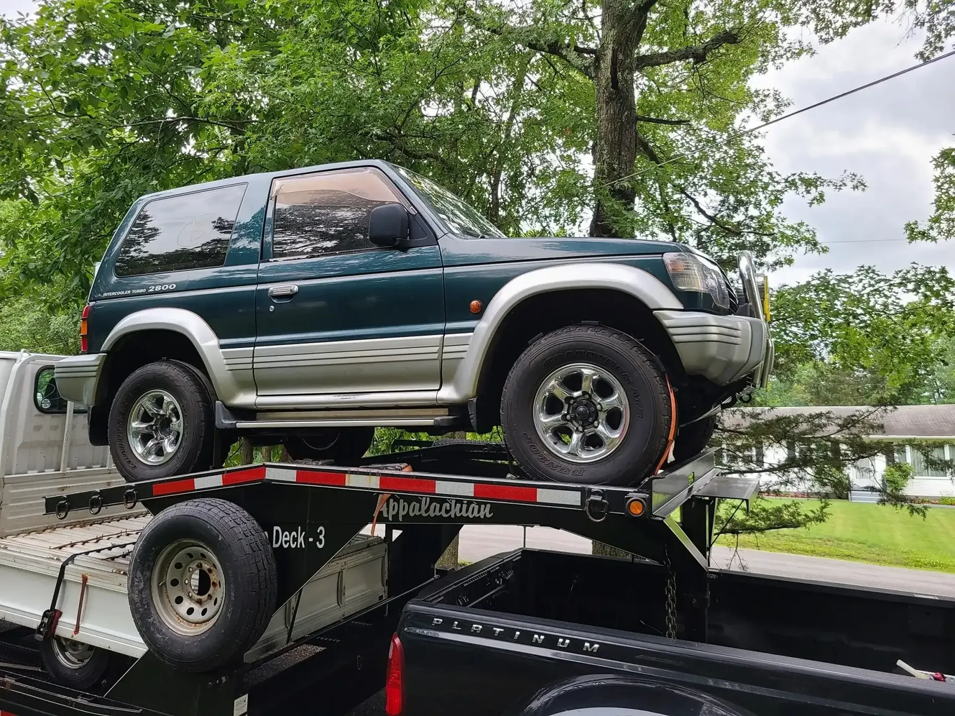 A dark green Mitsubishi Pajero SUV secured on a flatbed trailer towed by a black pickup truck.