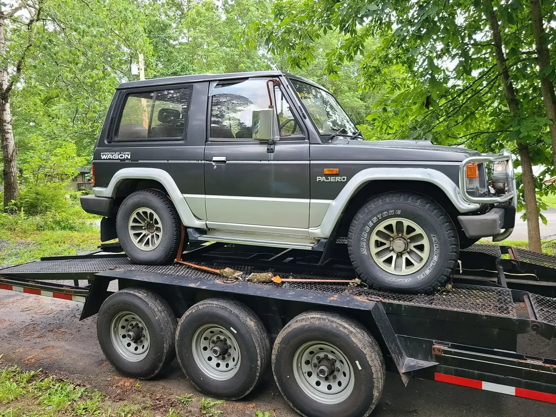 A matte black two-door Mitsubishi Pajero SUV secured on a triple-axle flatbed trailer outdoors amidst trees.