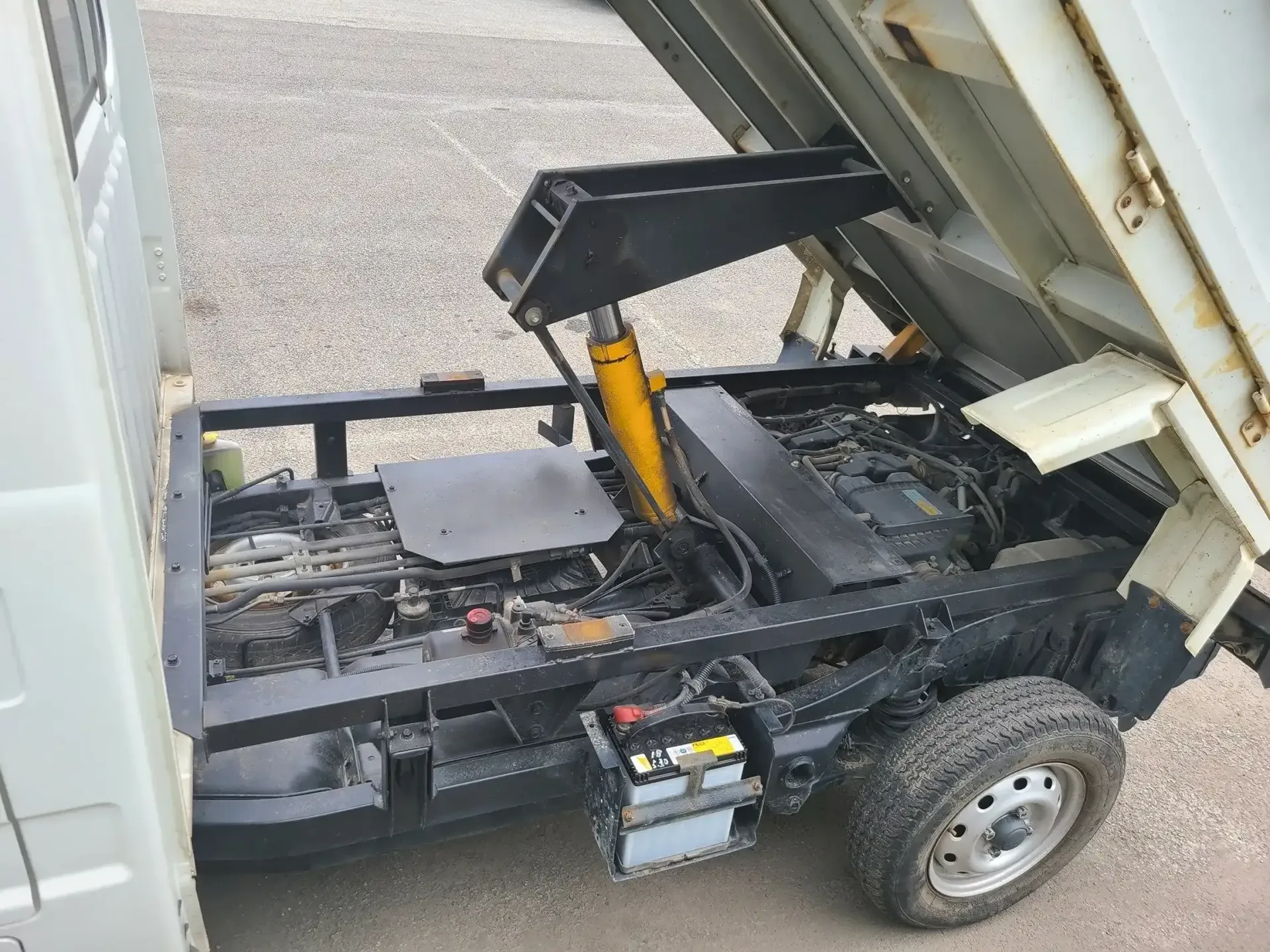 A white dump truck with its bed raised, revealing the black chassis, yellow hydraulic cylinder, engine, and battery.