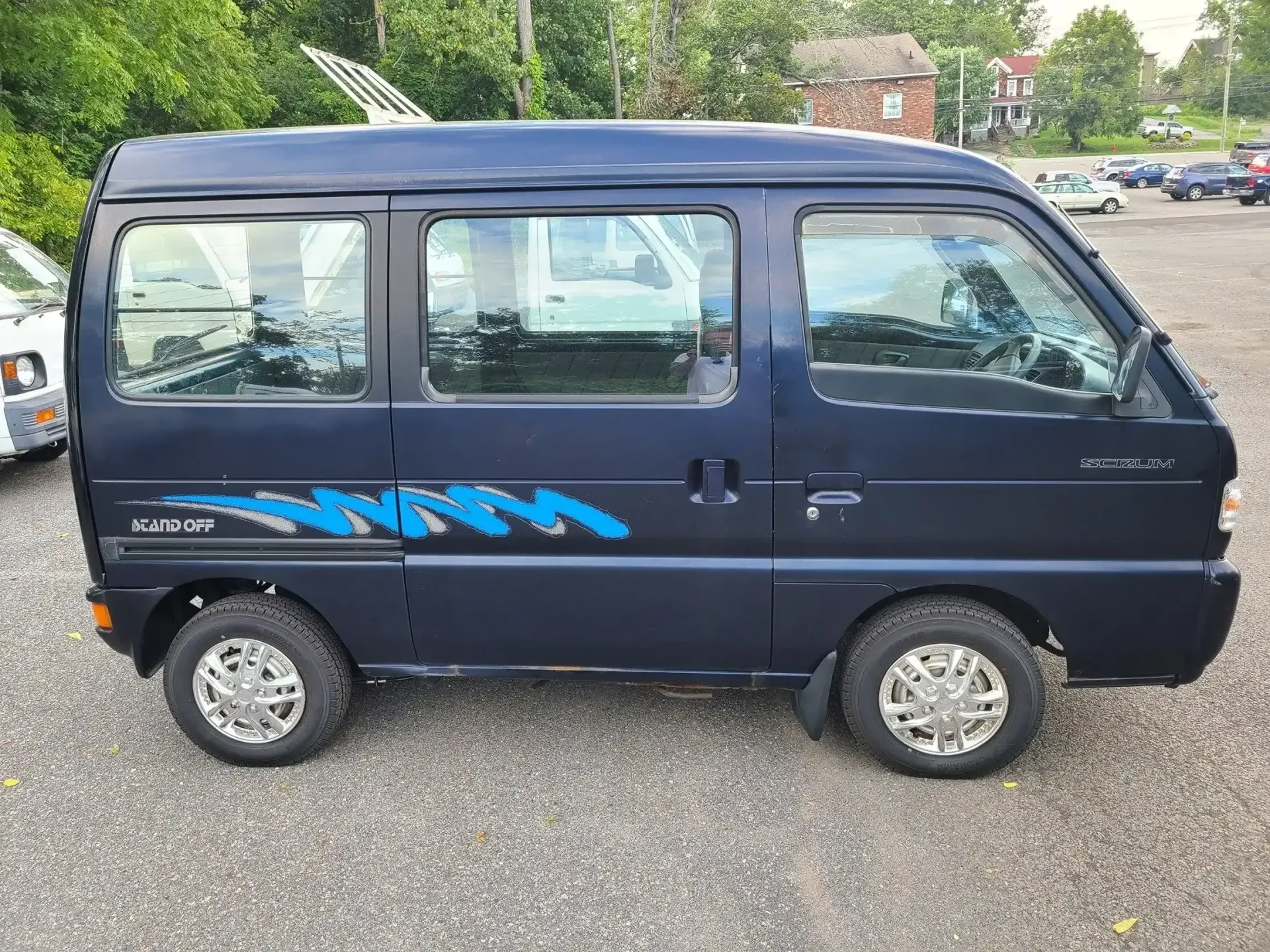 A dark blue Kei van parked on a gravel lot, featuring a blue and silver graphic design along the side.