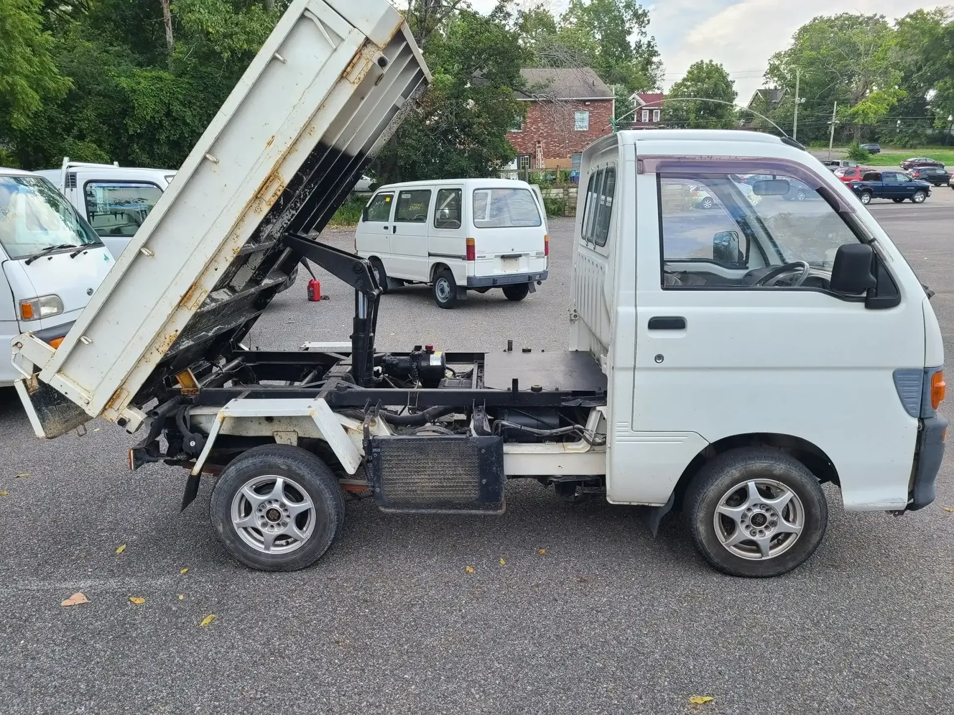 A white mini-dump truck parked on a gravel lot with its bed raised, surrounded by other small vehicles.