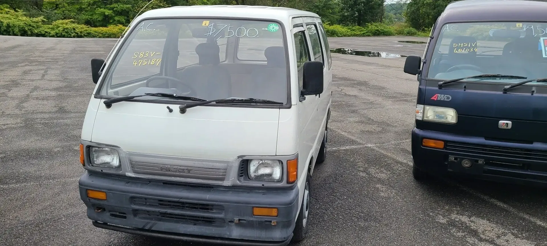 Two small, boxy vans, one white and one dark blue, parked on a gravel lot with a treeline in the background.