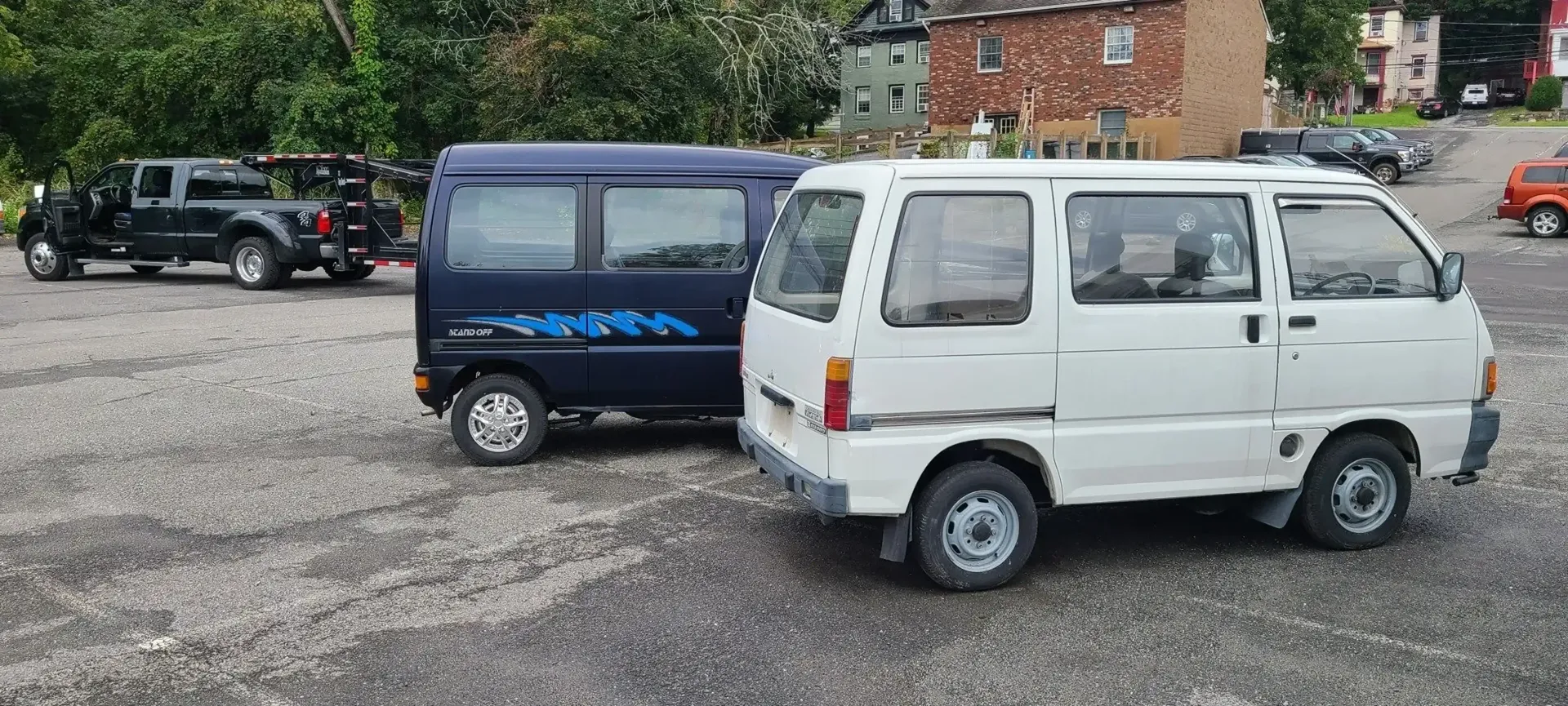 A blue and a white Japanese kei van parked on a gravel lot near a black truck and houses.
