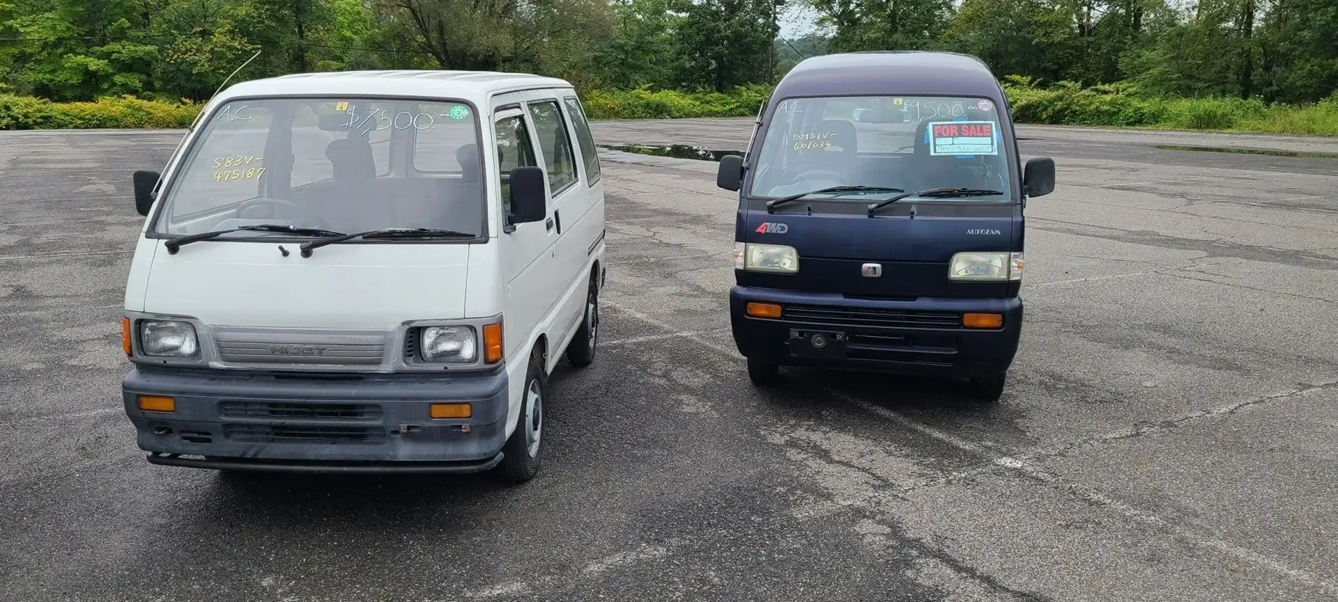A white and a dark blue Suzuki Carry van parked on a gravel lot with trees in the background.
