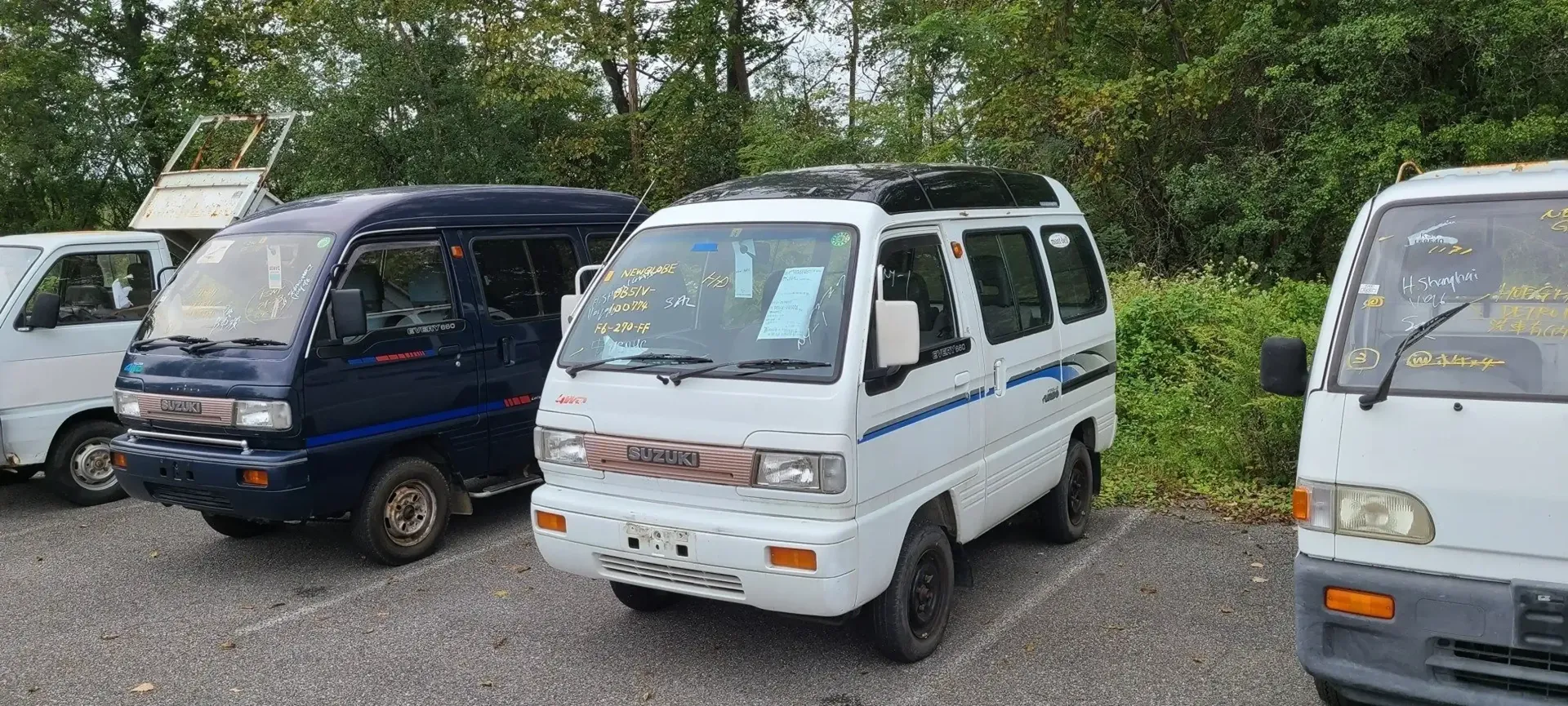 Three small, compact vans parked in a gravel lot with trees in the background.