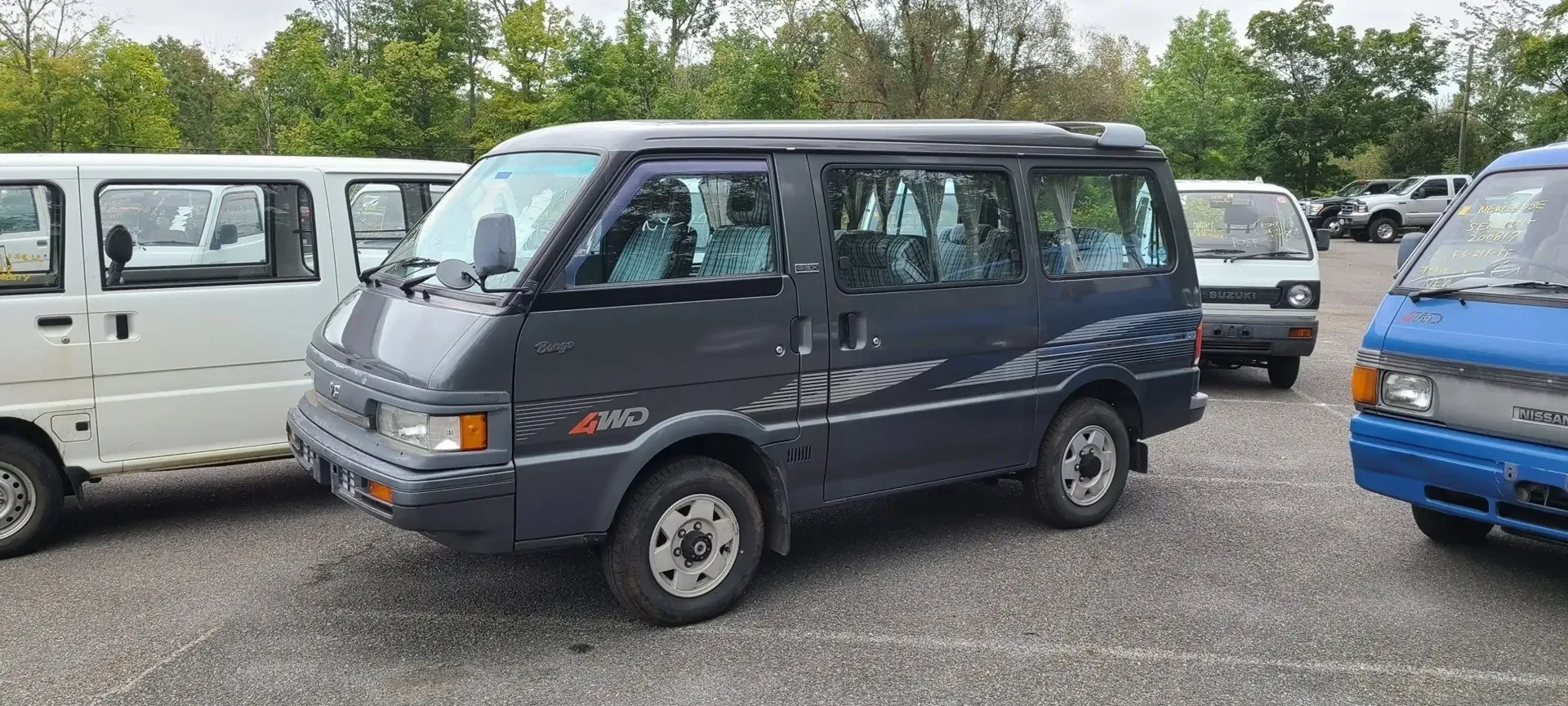 A dark grey 4WD Mitsubishi Delica van parked on a gravel lot among other similar Japanese microvans.