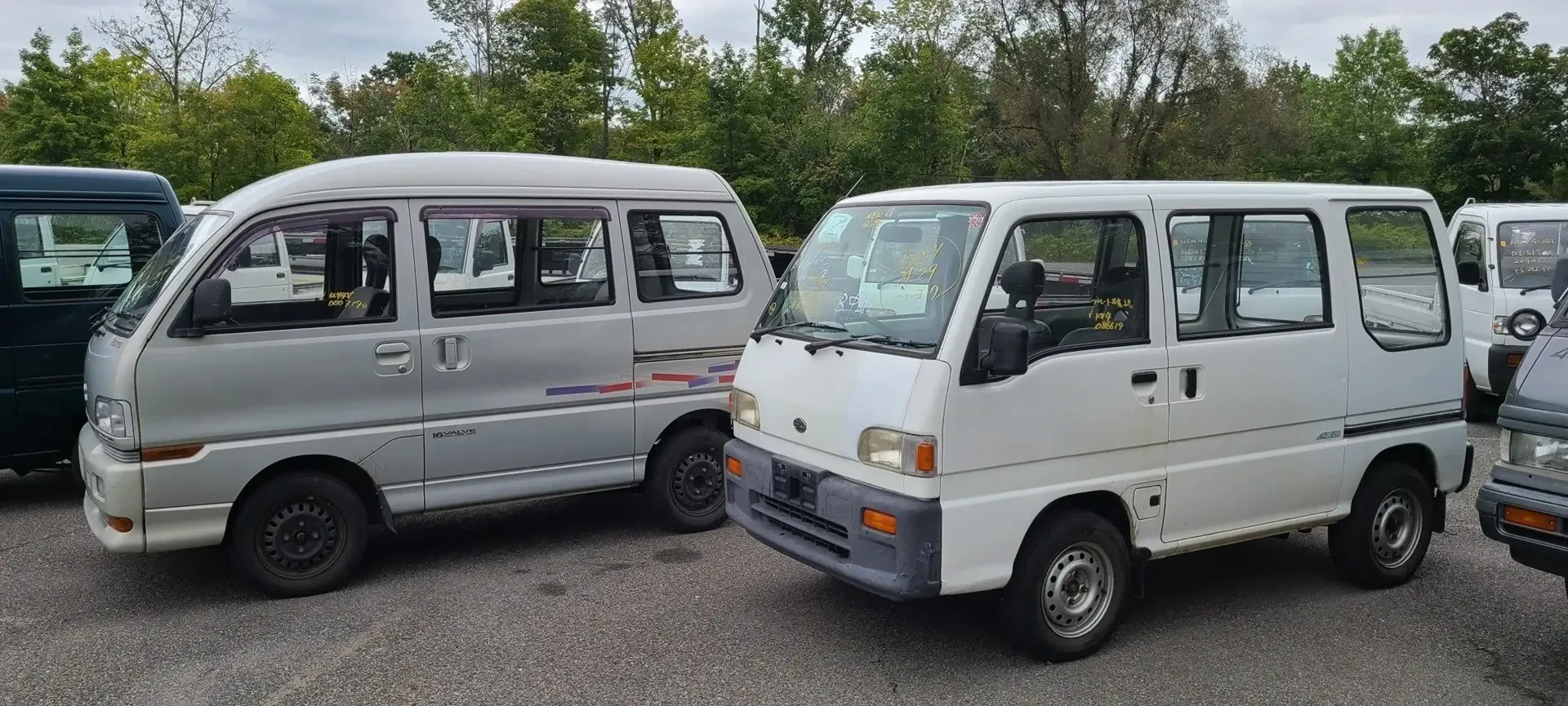 Two small boxy vans, one silver and one white, parked side-by-side on a gravel lot with trees in the background.