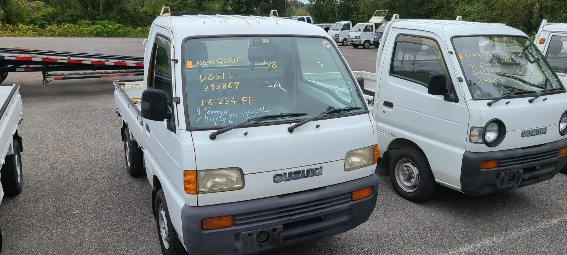 Two small white pickup trucks parked in a gravel lot with green trees in the background.