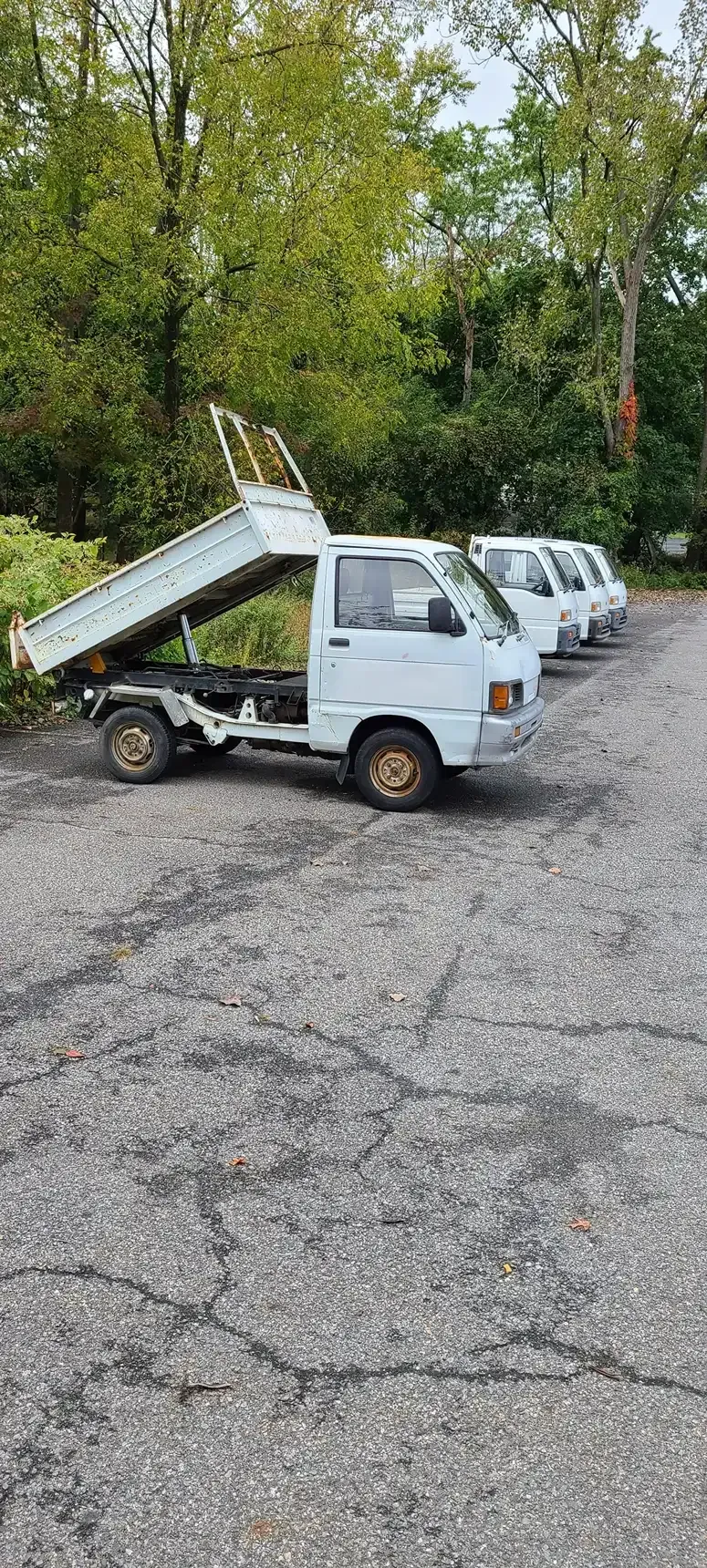 A row of white, small dump trucks parked on a gravel lot in front of trees, with the first truck's bed raised.