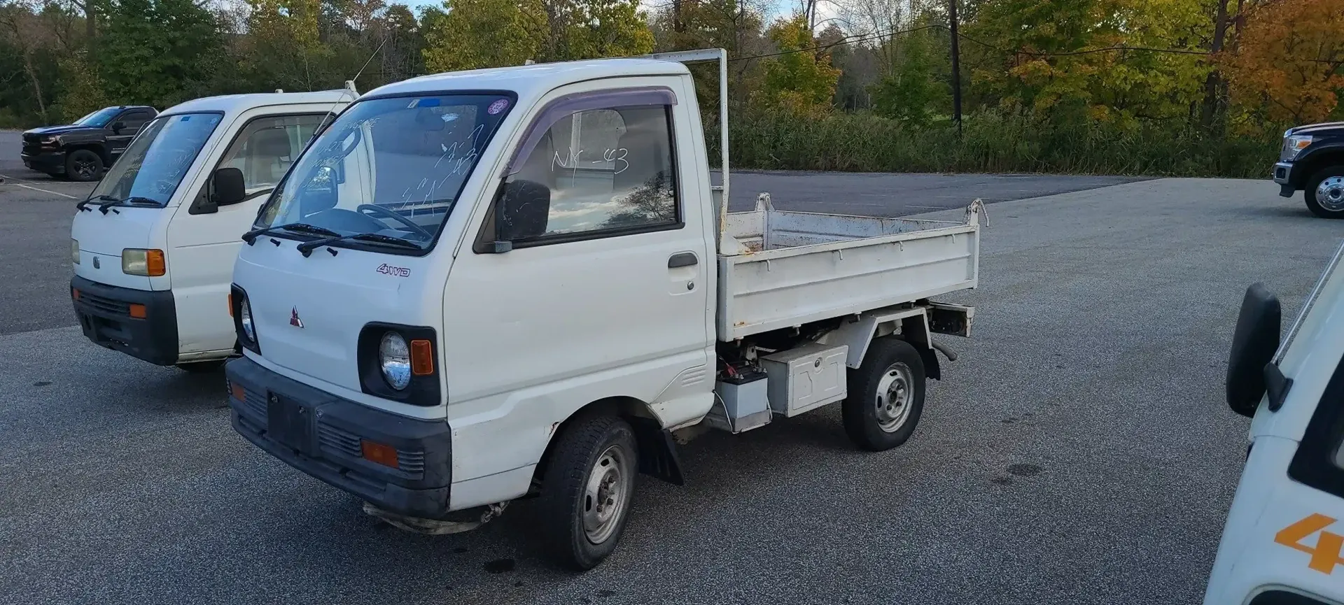 Several white kei trucks parked on a gravel lot during an autumn day.