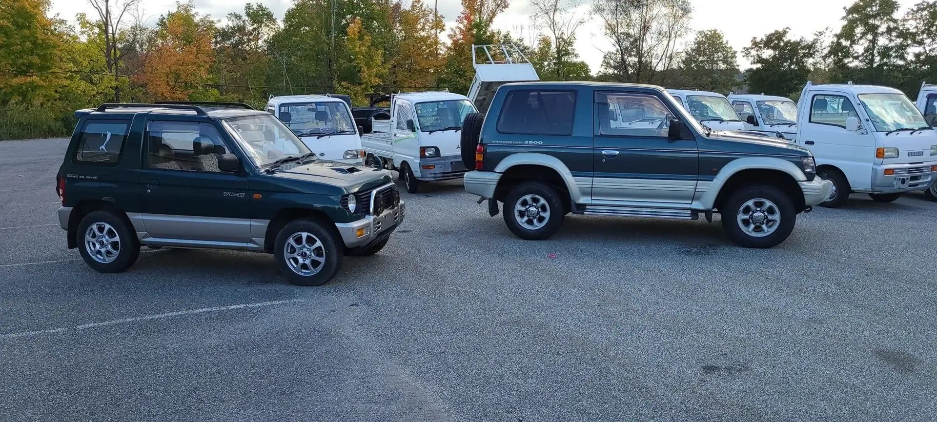 Two dark green Mitsubishi Pajero Mini SUVs parked on a gravel lot with small white trucks visible in the background.