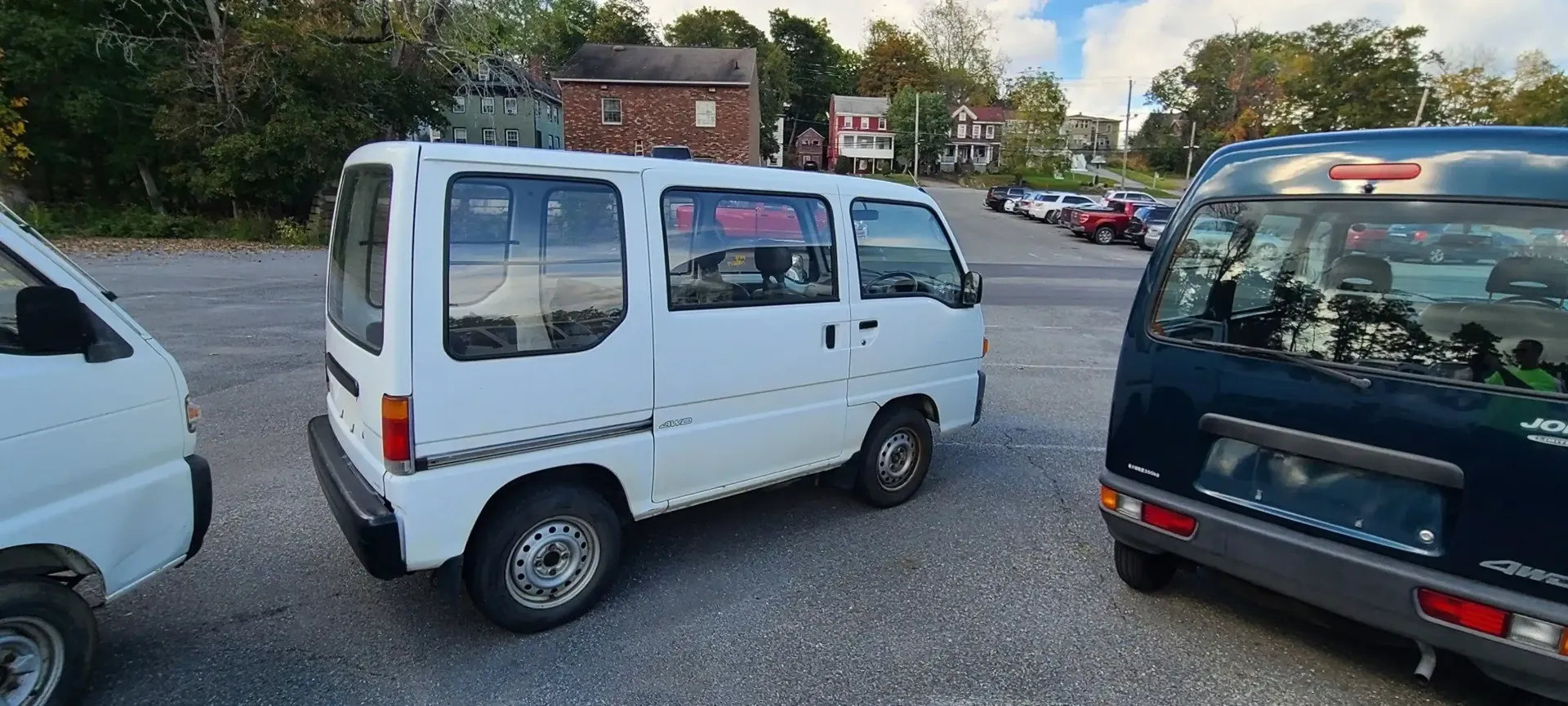 A small white kei van parked on a gravel lot next to a dark-colored vehicle.