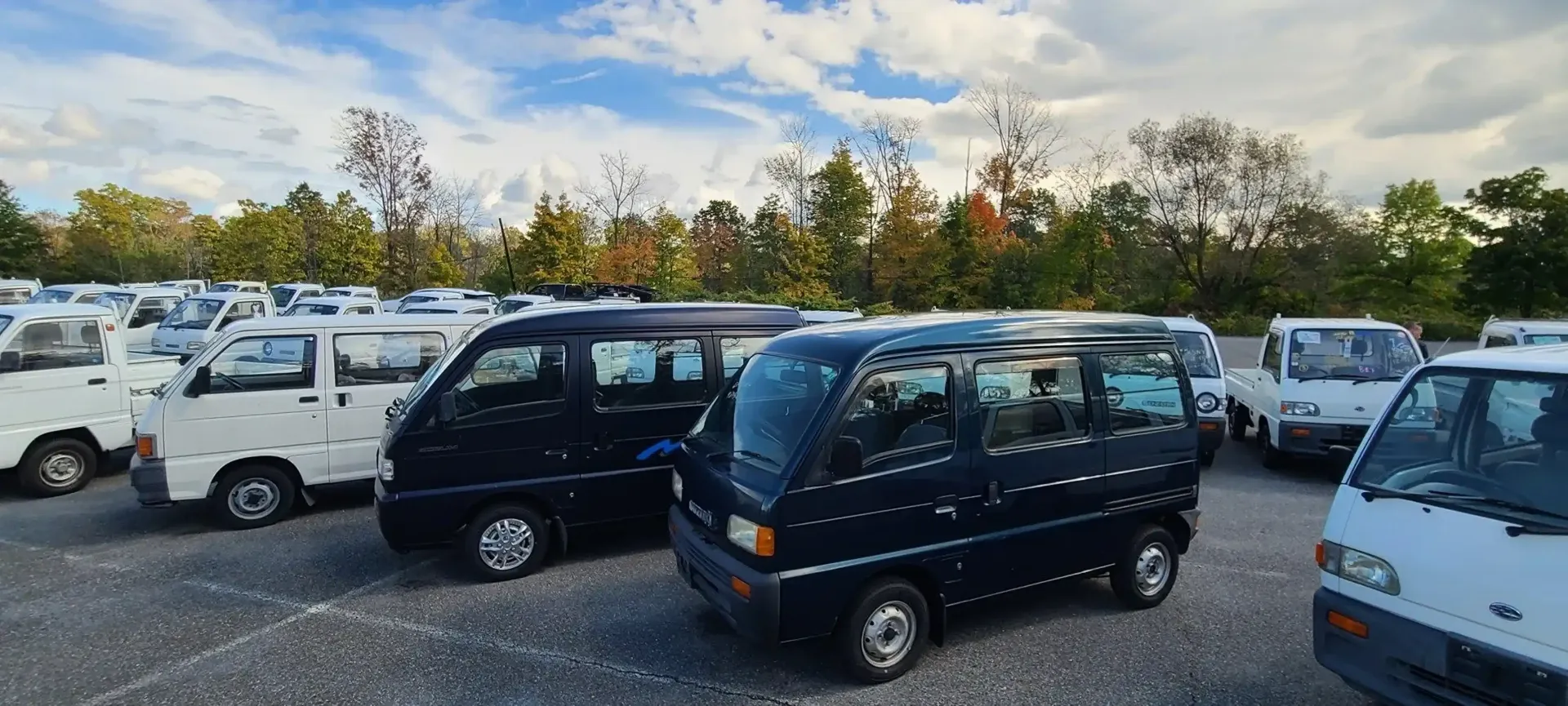 A parking lot filled with various white and dark-colored compact Kei vans and trucks under a blue sky with scattered clouds.