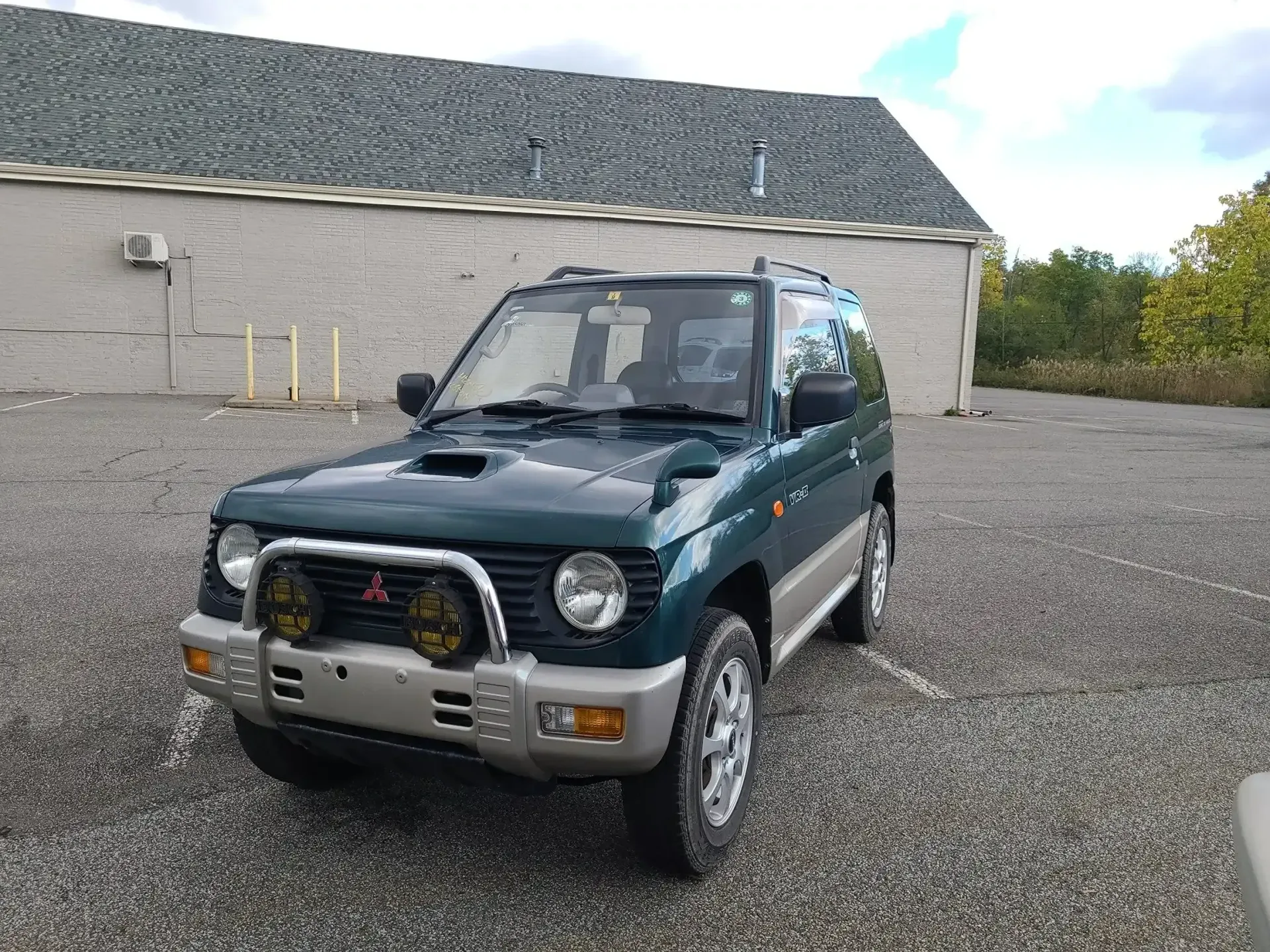 A green and silver Mitsubishi Pajero parked on a gravel lot in front of a building.