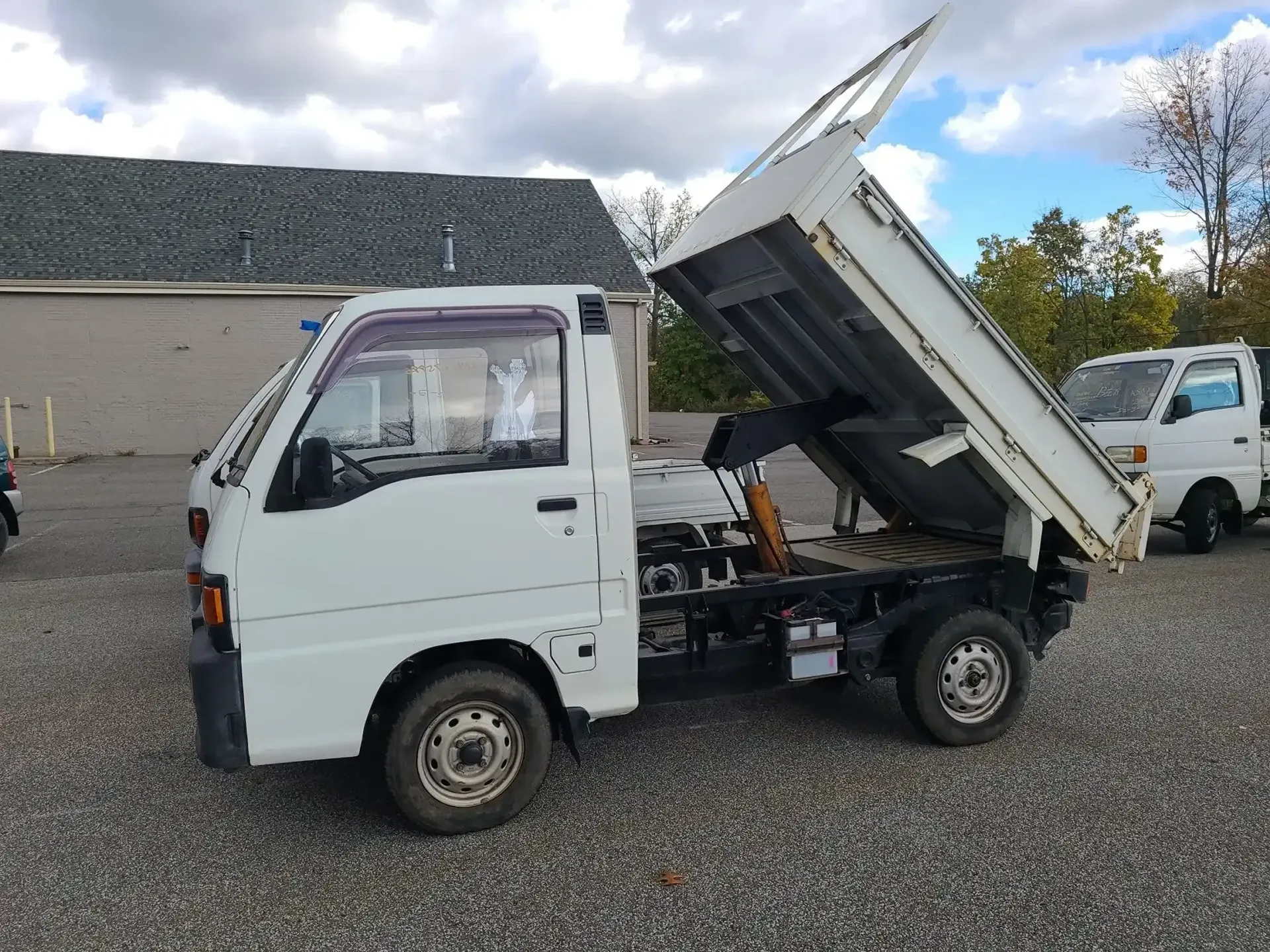 A white Kei dump truck with its bed raised, parked on a gravel lot under a cloudy sky.