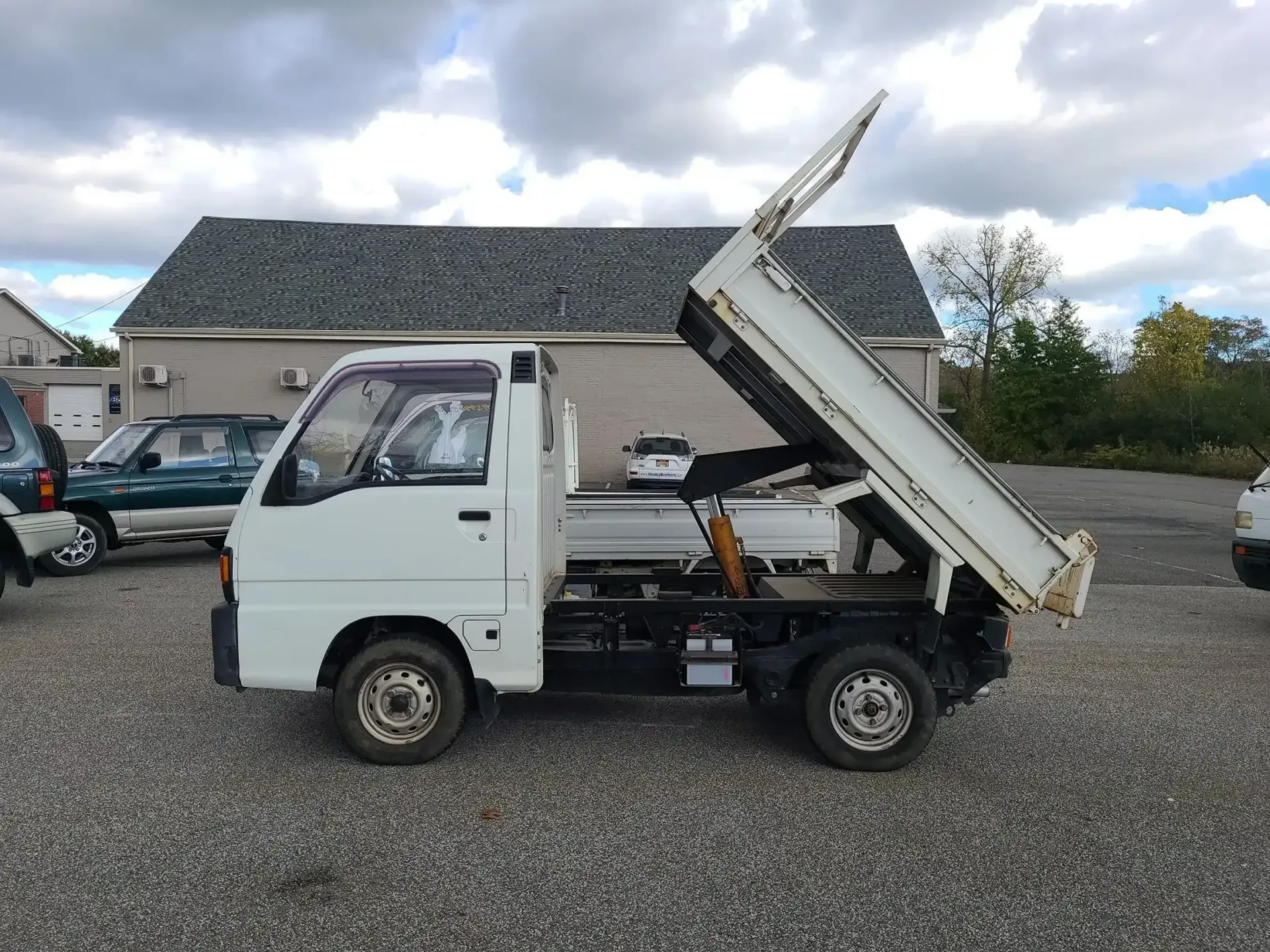 A white mini dump truck with its bed tilted upward, parked on a gravel lot in front of a building.
