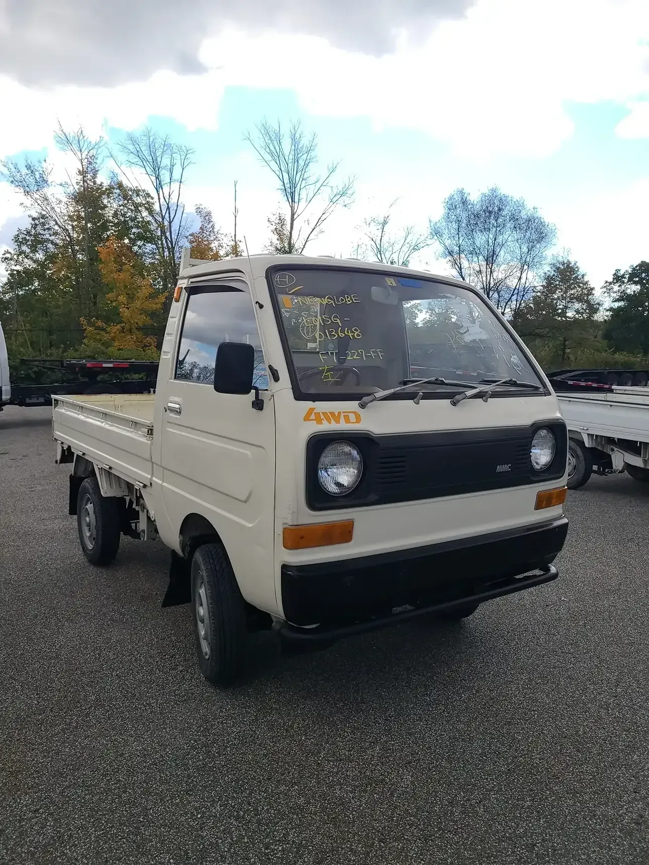 A beige, two-door kei truck parked on a gravel lot under a cloudy sky.