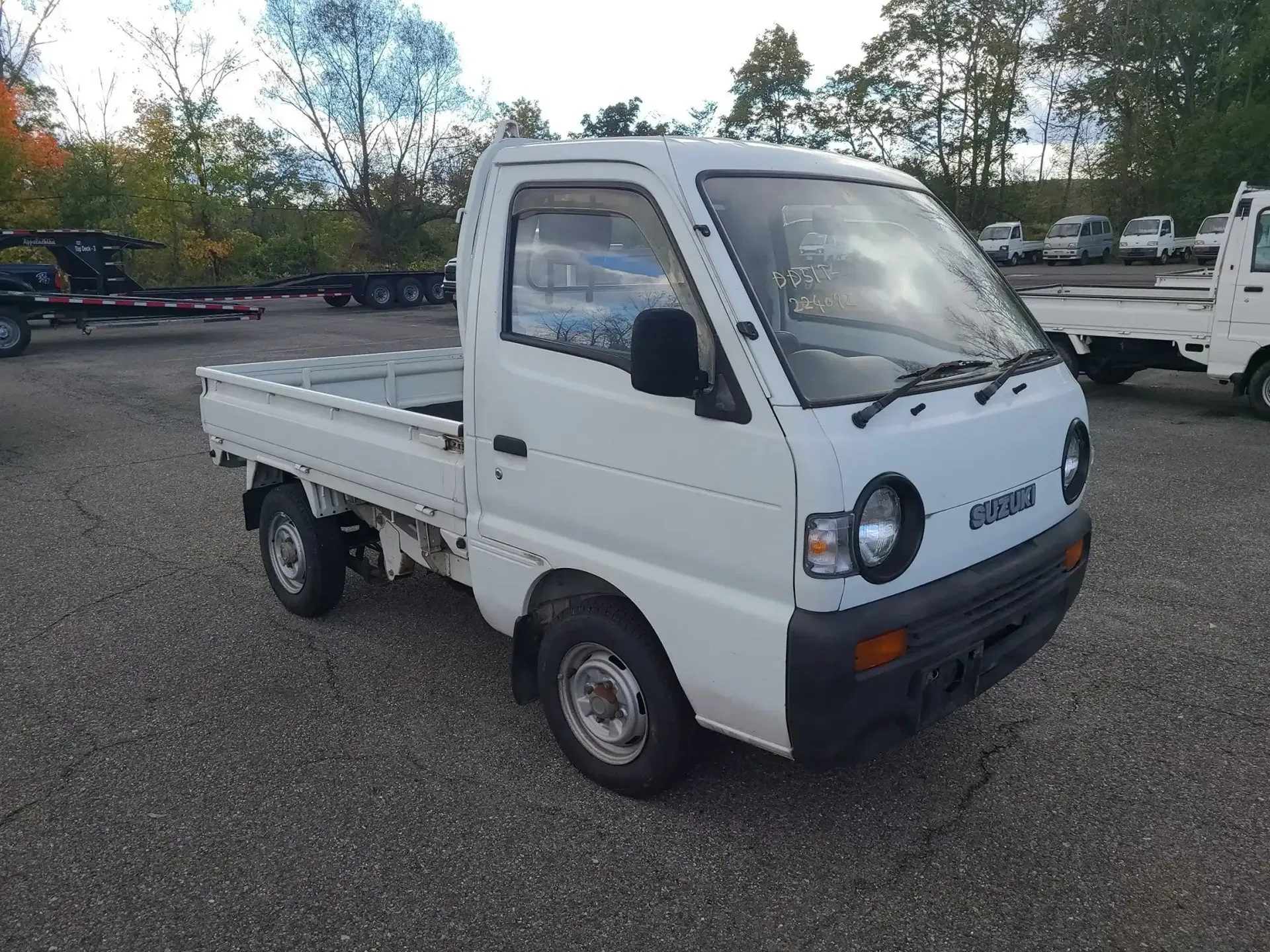 A white Suzuki Carry kei truck parked on a gravel lot, with another white truck partially visible in the background.