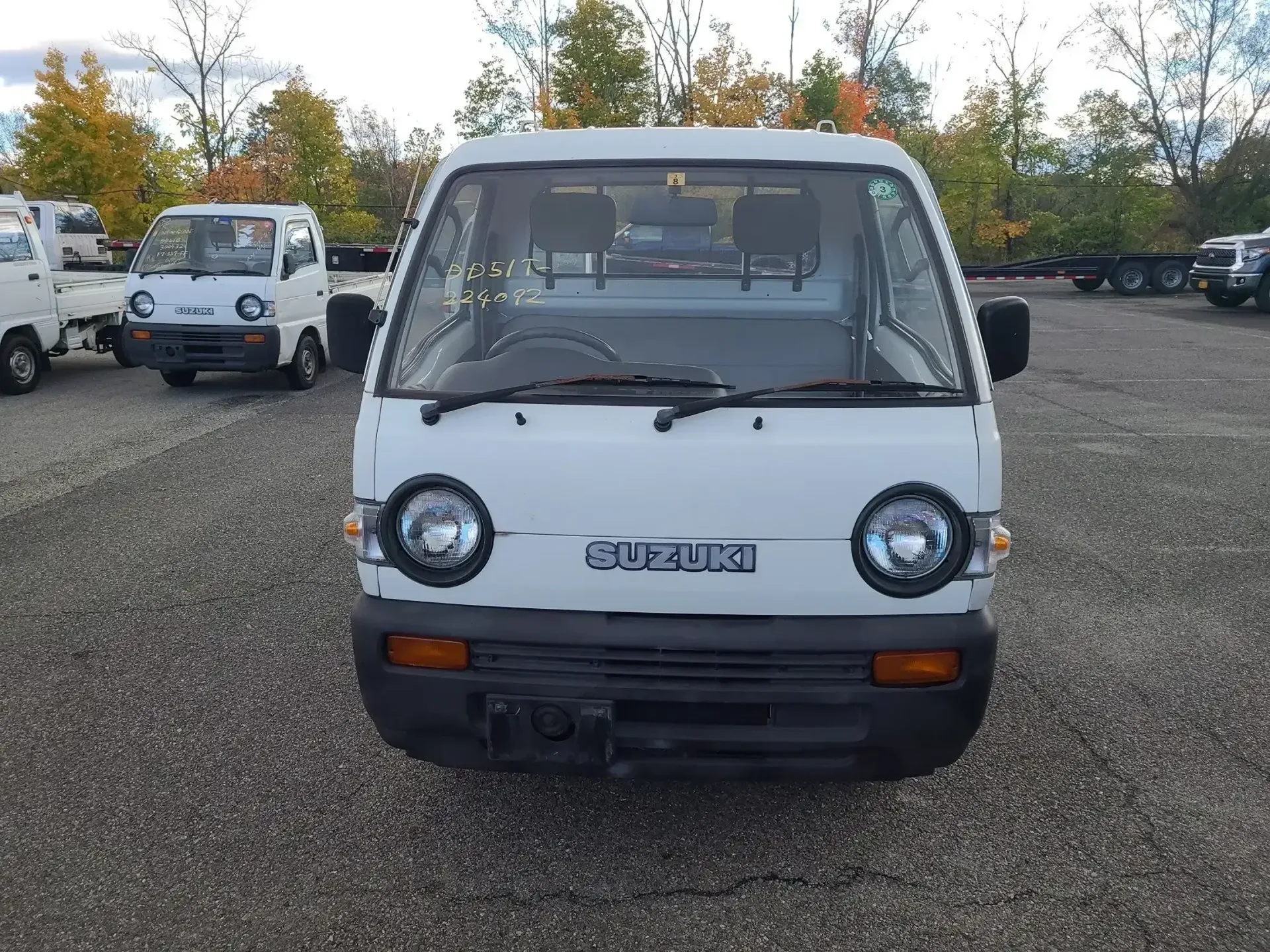 A white Suzuki Carry kei truck sits in a gravel lot, viewed from the front with another similar truck visible behind.