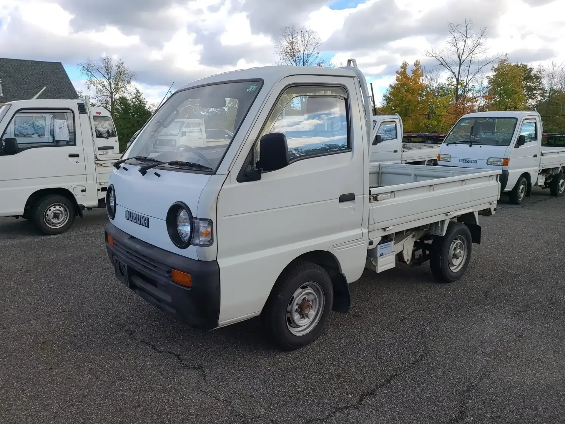 A white kei truck parked on a gravel lot under a cloudy sky, with other similar trucks visible in the background.