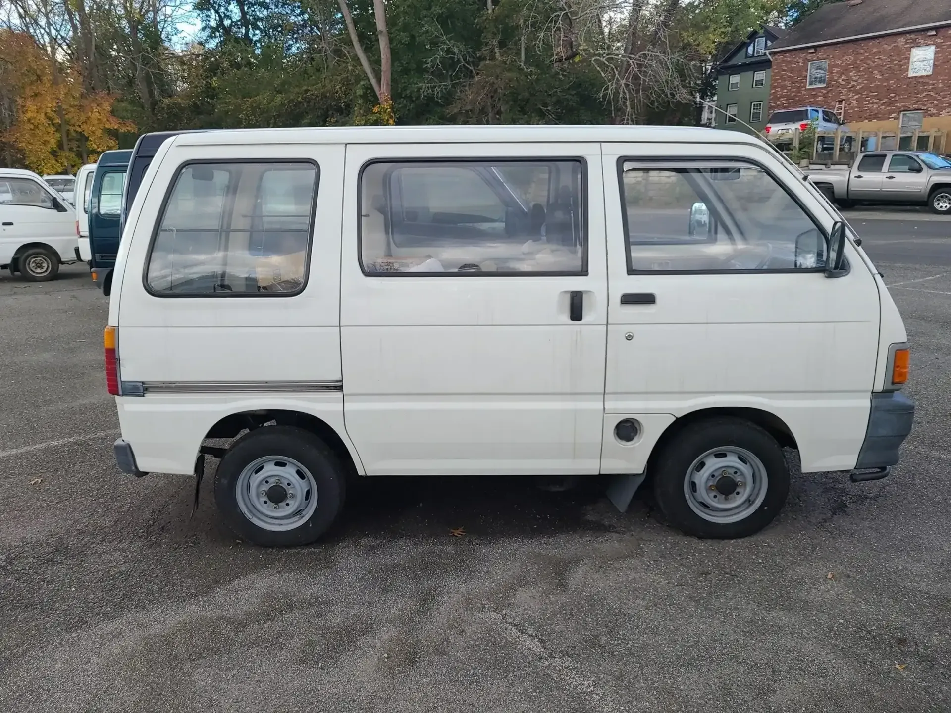 A white, boxy compact van parked on a gravel lot with other vehicles visible in the background.
