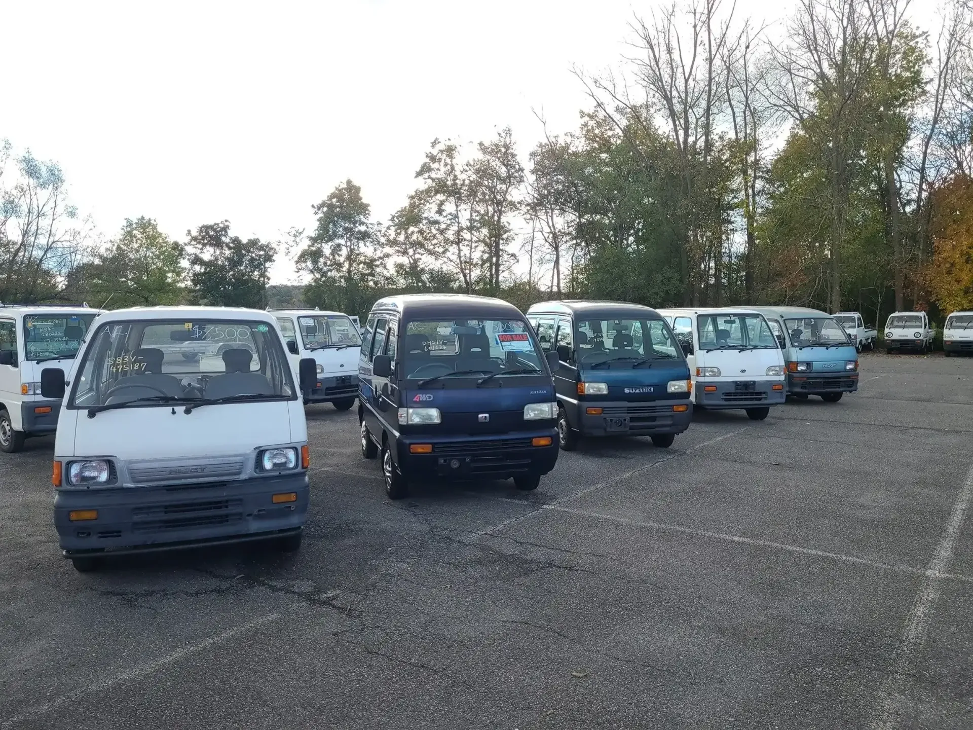 A row of small, boxy kei trucks parked on a gravel lot under a bright, overcast sky.