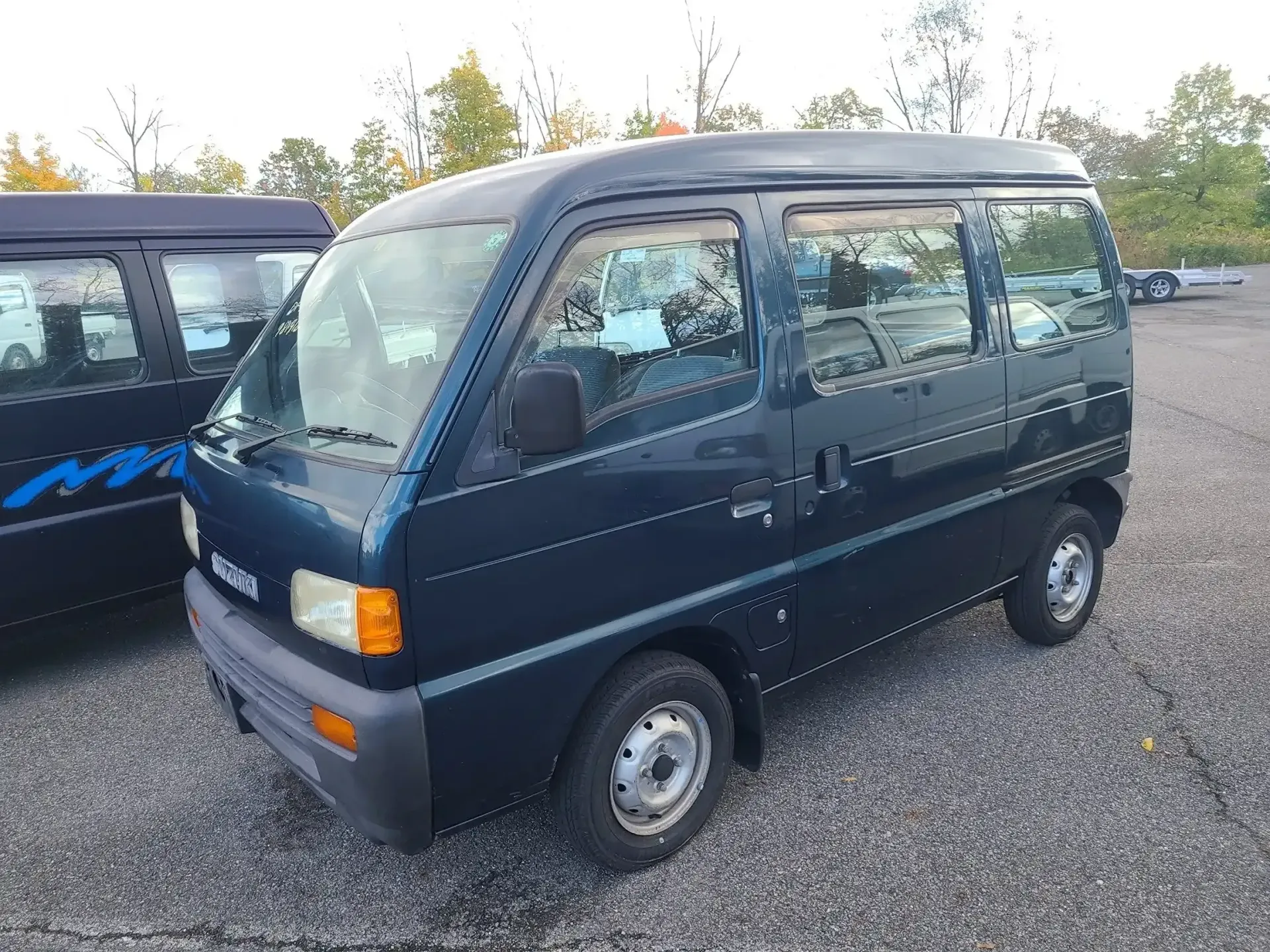 A dark blue, compact Suzuki Kei van parked on a gravel lot next to another similar vehicle.