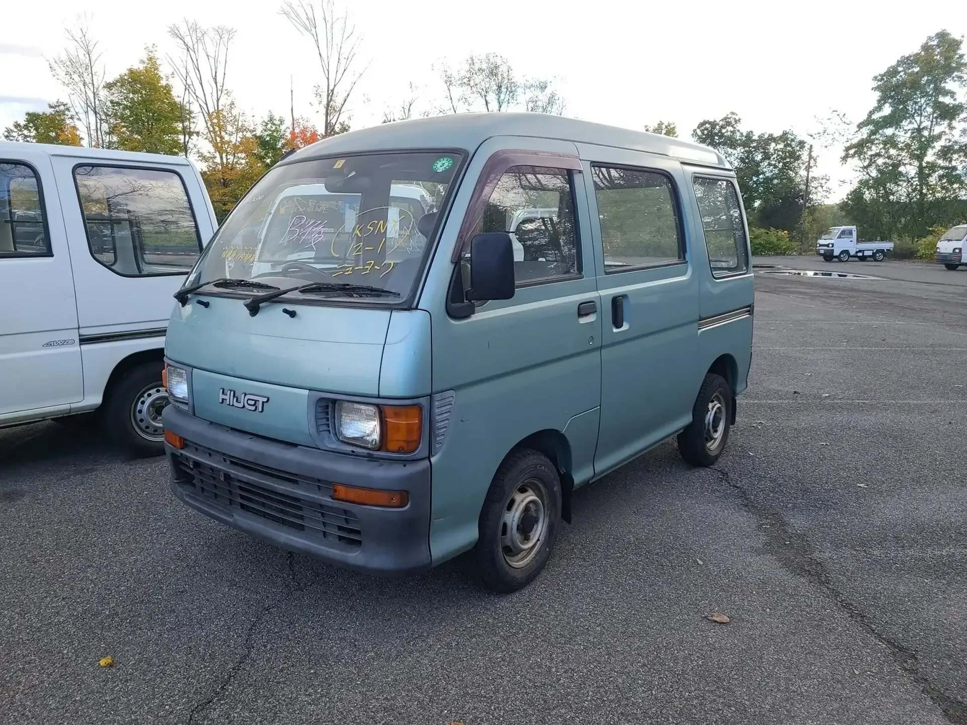 A light blue, boxy Japanese kei van parked on a gravel lot, with a white van partially visible on the left.