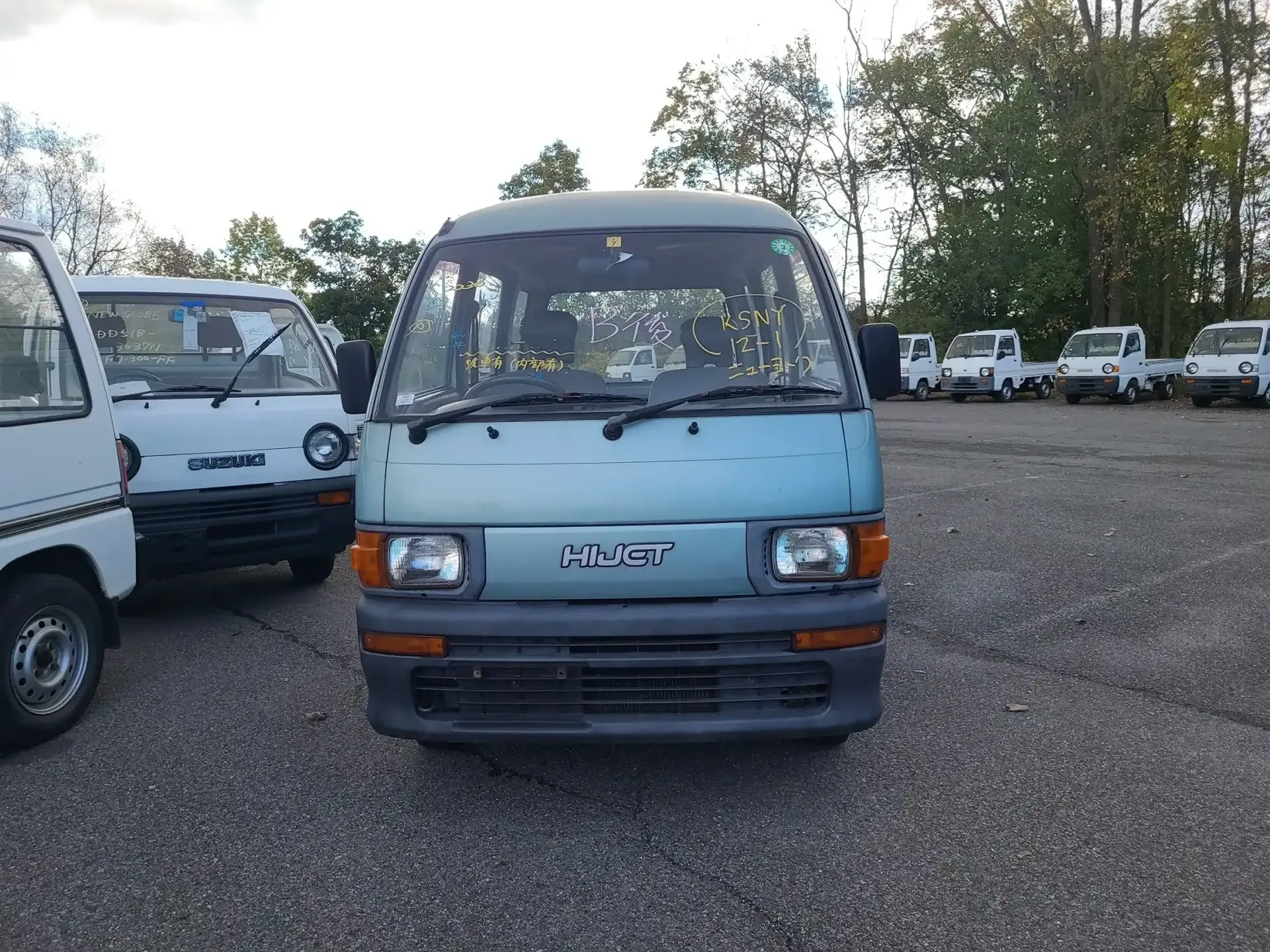A light blue Daihatsu Hijet van parked in a gravel lot among other similar small commercial vehicles.