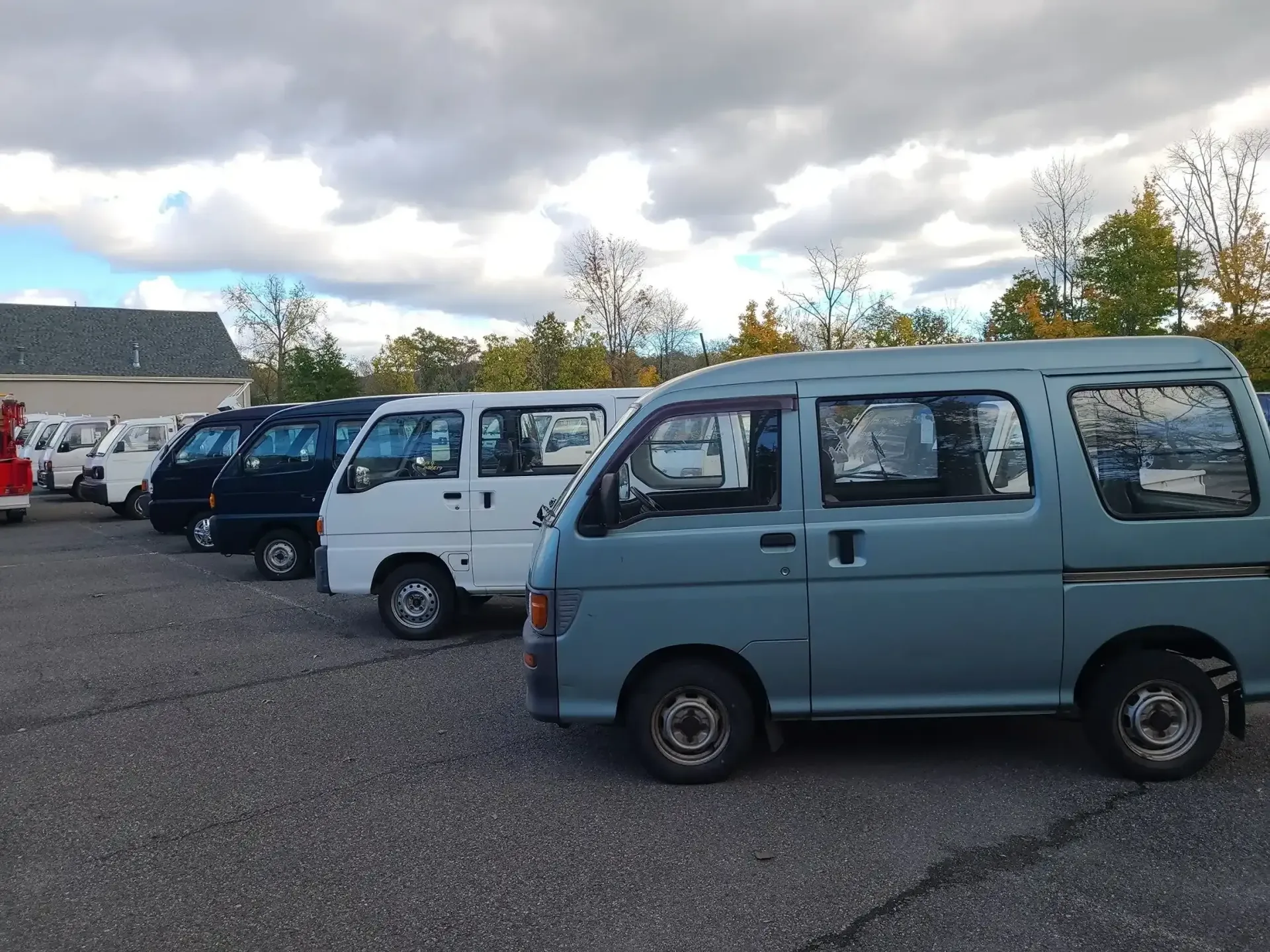 A line of small, light-colored box vans parked on a gravel lot under a cloudy sky.