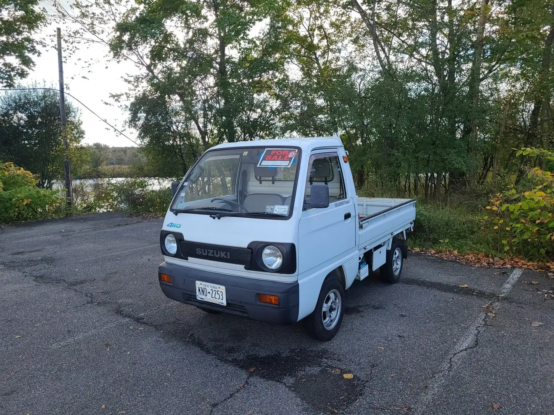 A white Suzuki Carry kei truck parked on a gravel lot with trees in the background.