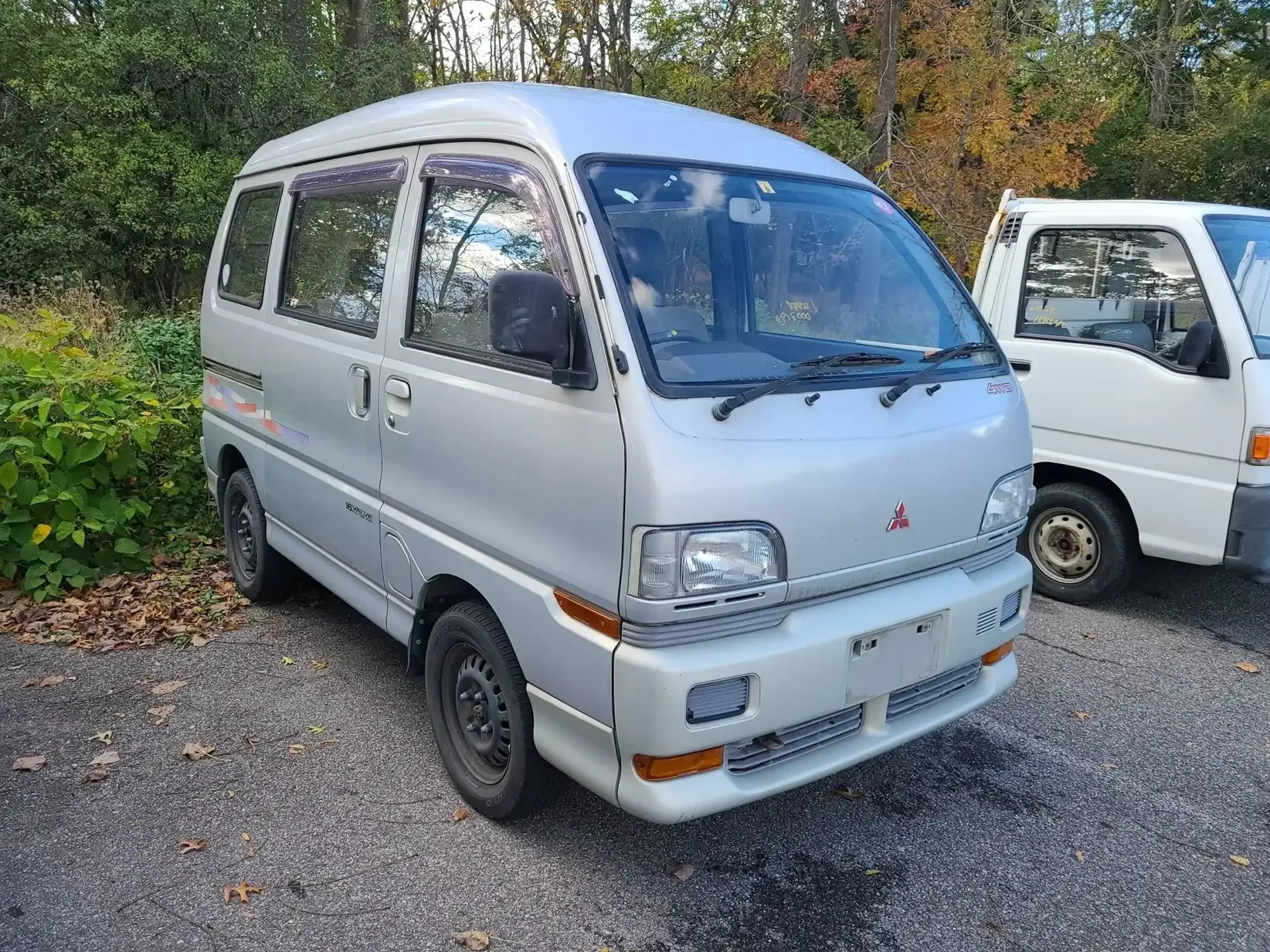 A silver Mitsubishi kei van parked on a gravel lot next to a white truck with trees in the background.