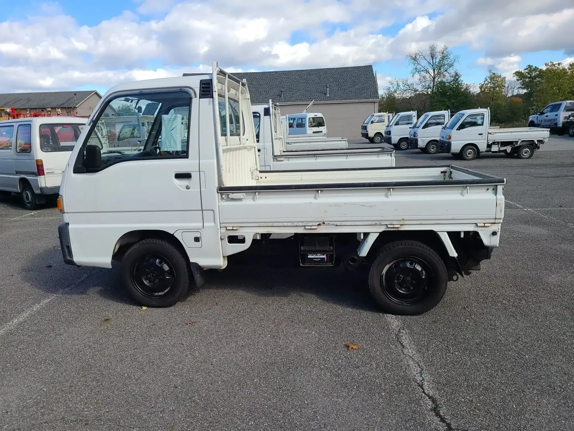 A white Kei truck parked on a gravel lot under a cloudy sky, with several other similar trucks in the background.
