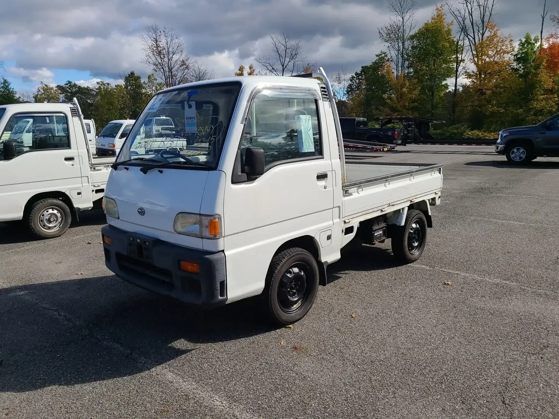 A white, compact kei truck parked on a gravel lot under a partly cloudy sky, with another truck partially visible nearby.