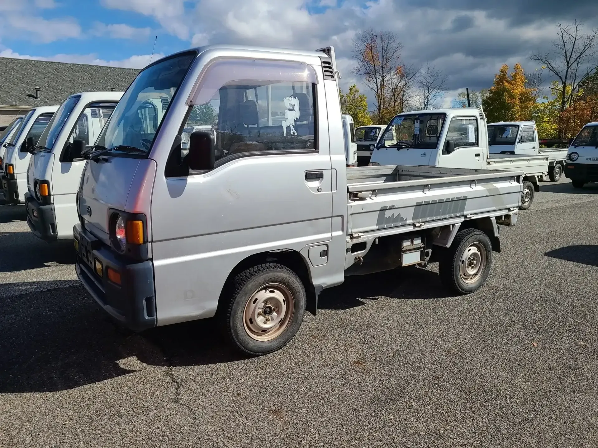 A silver kei truck parked in a gravel lot filled with other similar small utility vehicles under a cloudy sky.
