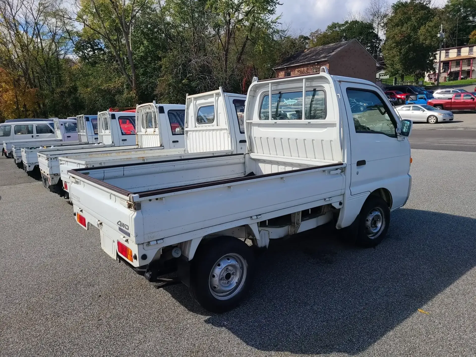 A row of small, white Kei trucks parked on a gravel lot under a bright, sunny sky.