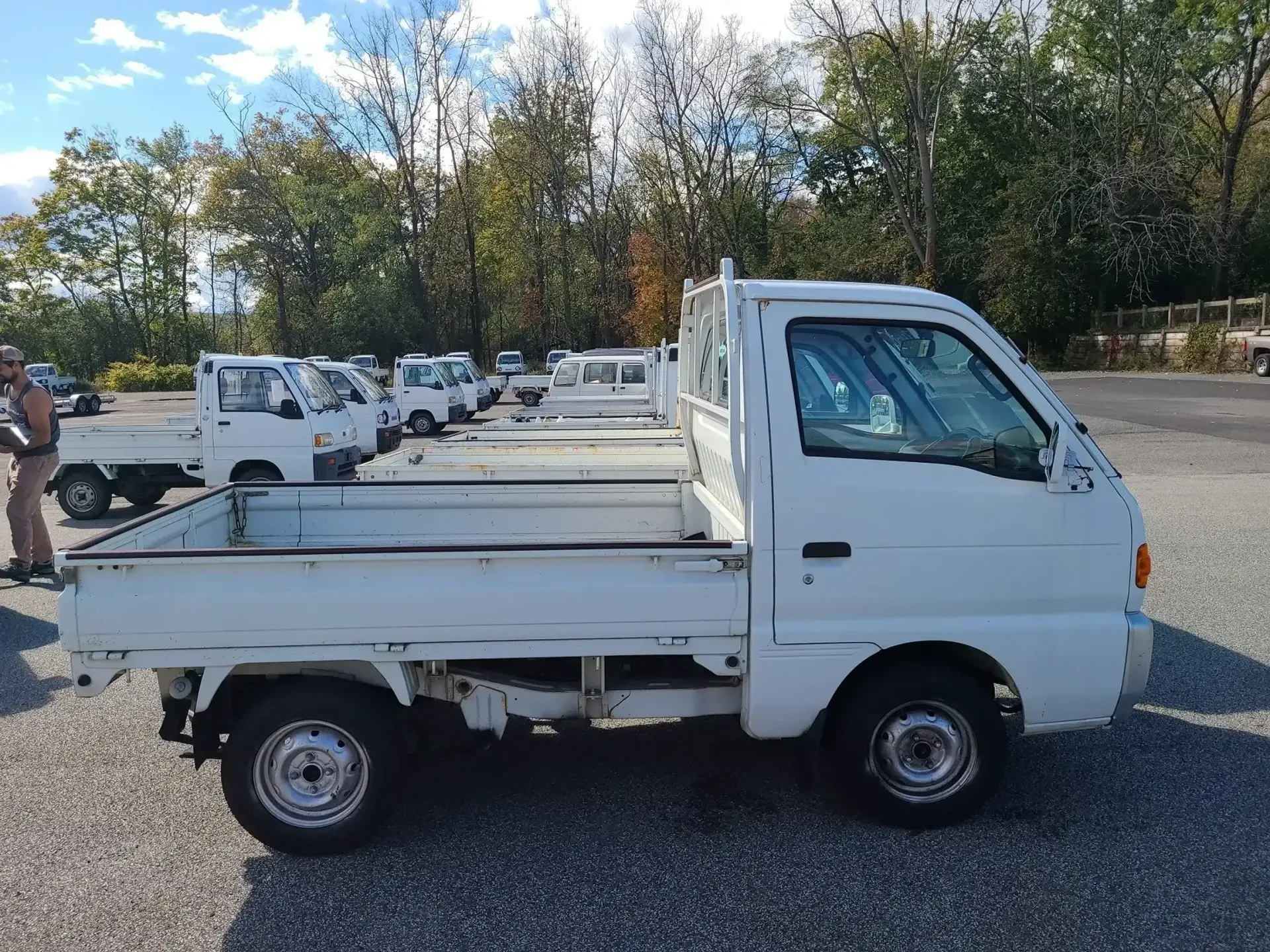 A white Kei truck parked in a gravel lot with a line of identical trucks visible in the background.