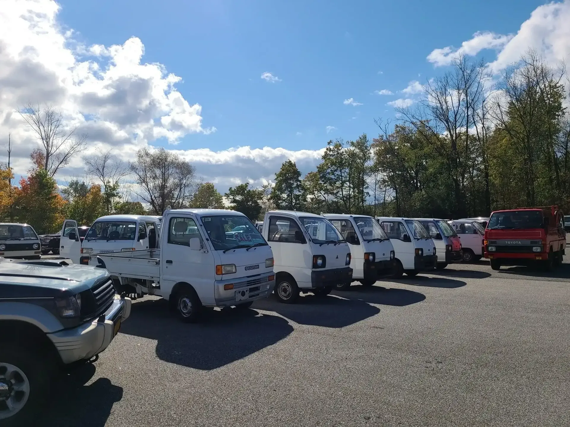 A row of white compact trucks parked on a gravel lot under a sunny, cloudy sky, with a red truck at the end.