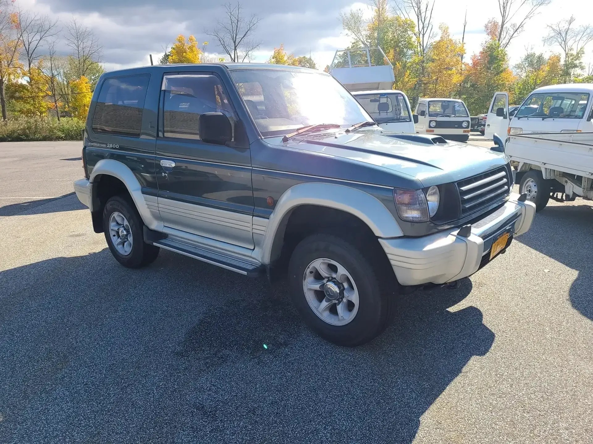 A dark green and silver two-door Mitsubishi Pajero SUV parked on a gravel lot under a bright, sunny sky.